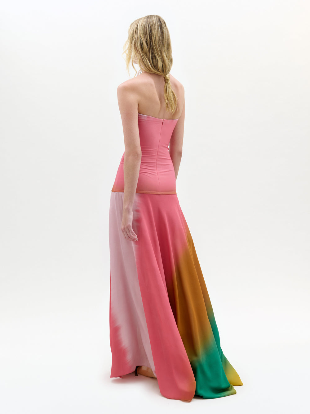 Una persona con el pelo largo, rubio y ondulado mira de frente, luciendo el vestido rosa sin tirantes Inari -un maxi columna hasta el suelo con secciones rosas, naranjas y verdes- sobre un fondo blanco liso.