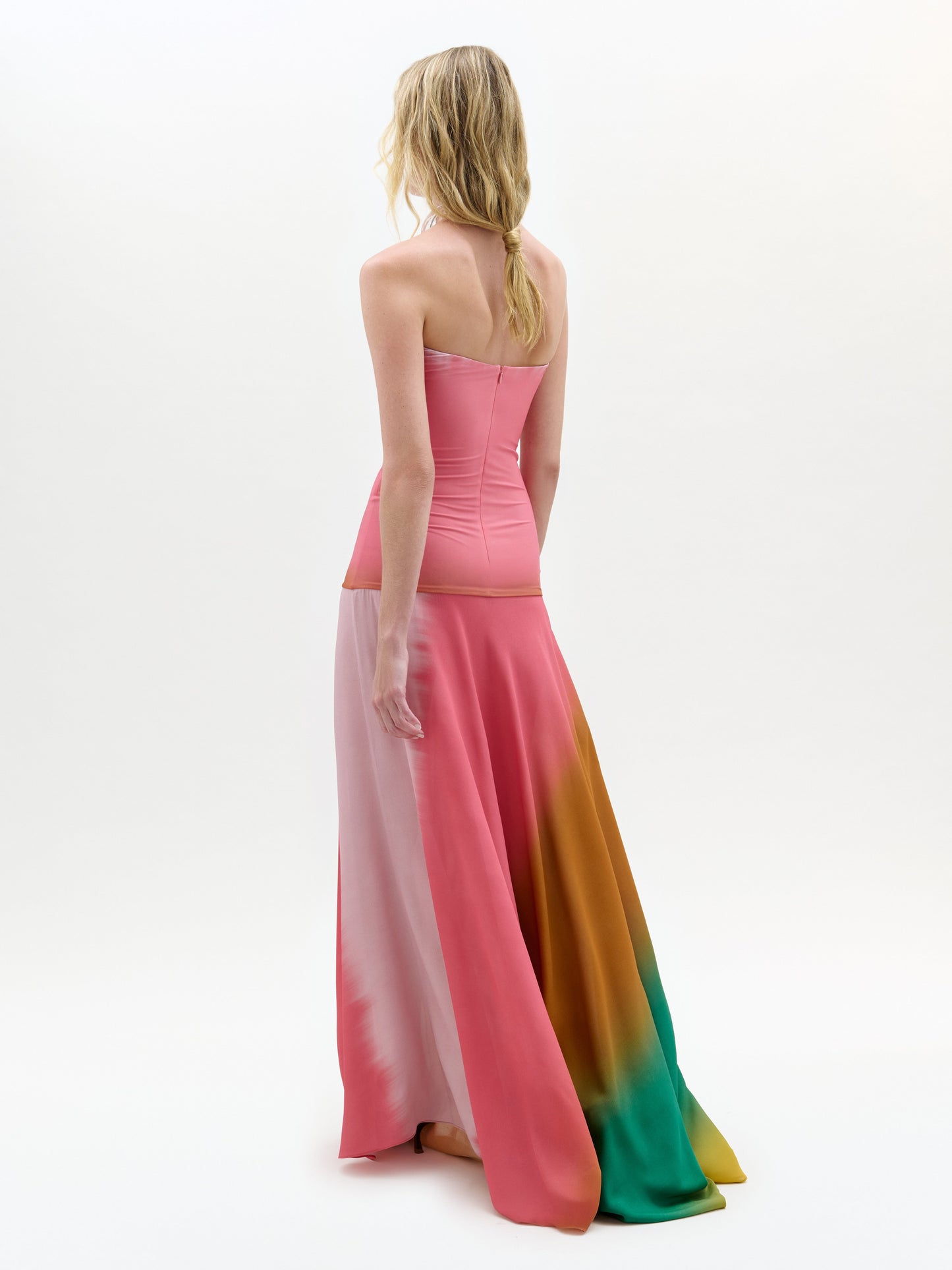 Una persona con el pelo largo, rubio y ondulado mira de frente, luciendo el vestido rosa sin tirantes Inari -un maxi columna hasta el suelo con secciones rosas, naranjas y verdes- sobre un fondo blanco liso.