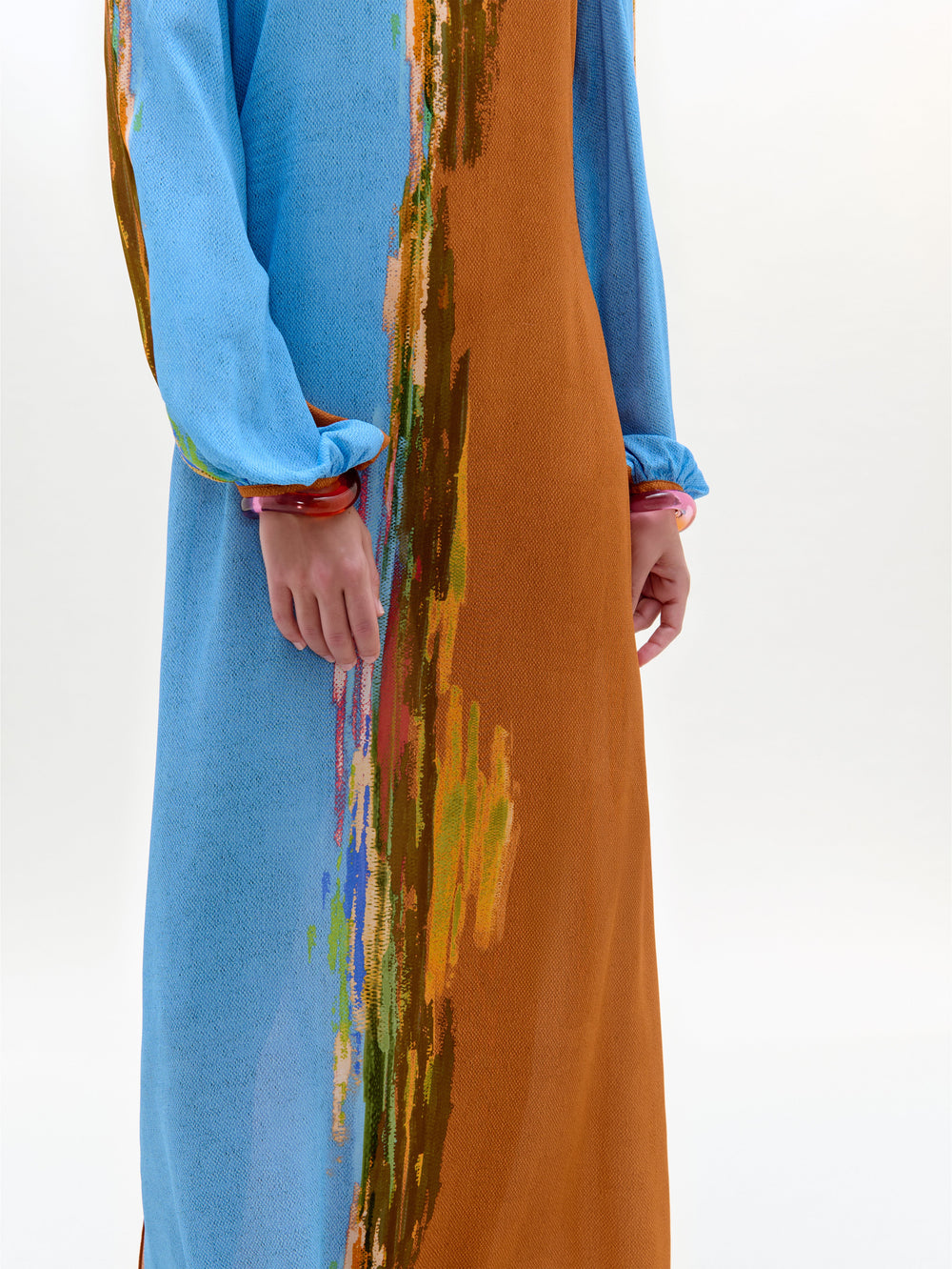 Una persona lleva el vestido Janys, un maxi con mangas y costados azules, un panel naranja-marrón y una raya vertical multicolor abstracta. Sólo se ven el torso y los brazos sobre un fondo liso. Prenda hecha en Colombia.