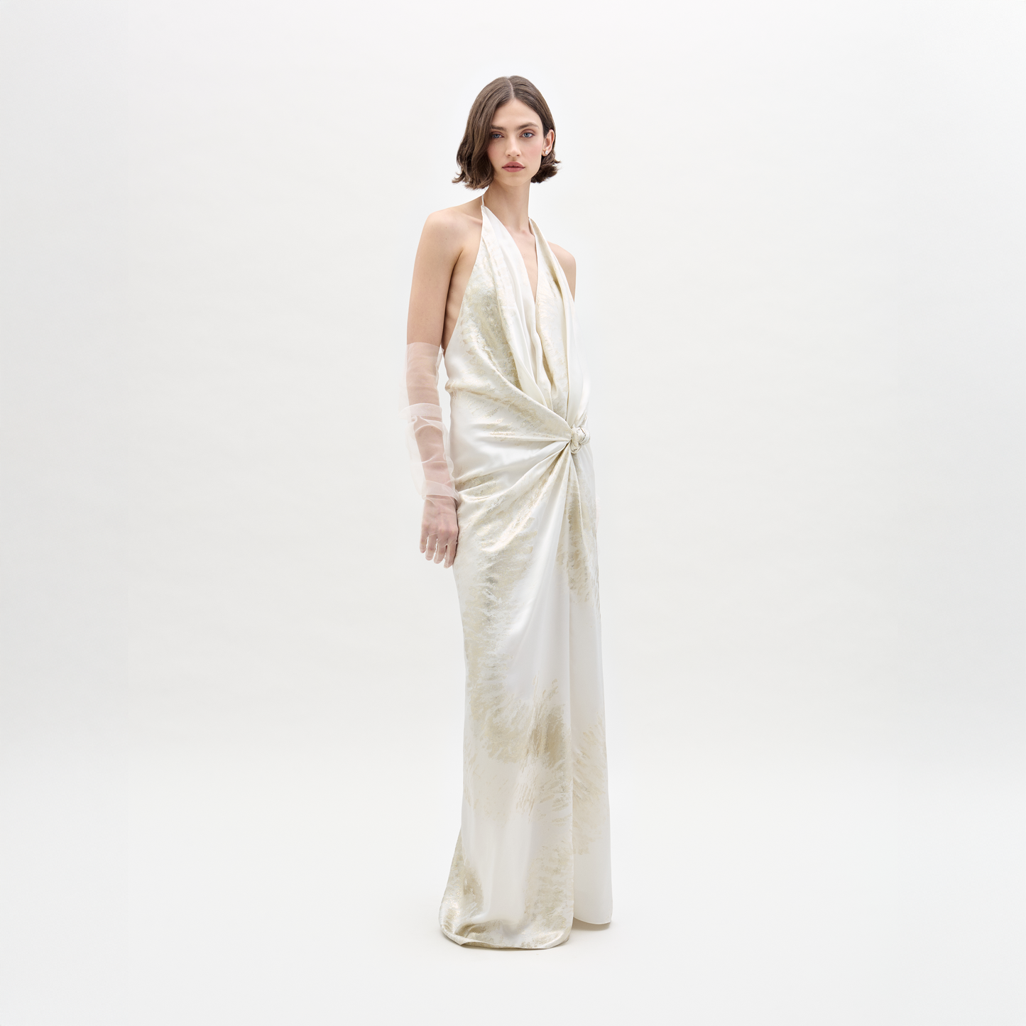 Una mujer con cabello castaño corto y expresión neutra posa sobre un fondo blanco con el Vestido Jazmin, un vestido largo sin mangas de color marfil con escote halter, detalle de nudo drapeado en la cintura y guantes transparentes.