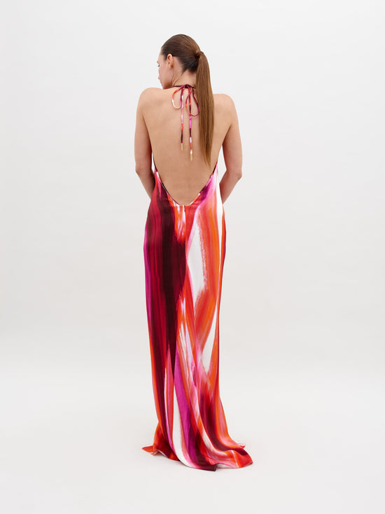 A woman stands against a plain background, wearing a colorful, flowing halter-neck Jazmin Dress Magenta Abstract Waves with a prominent knot detail at the waist. Available for pre-order now, the dress will ship by November 15th, 2024. She has her hands behind her back and is looking down.