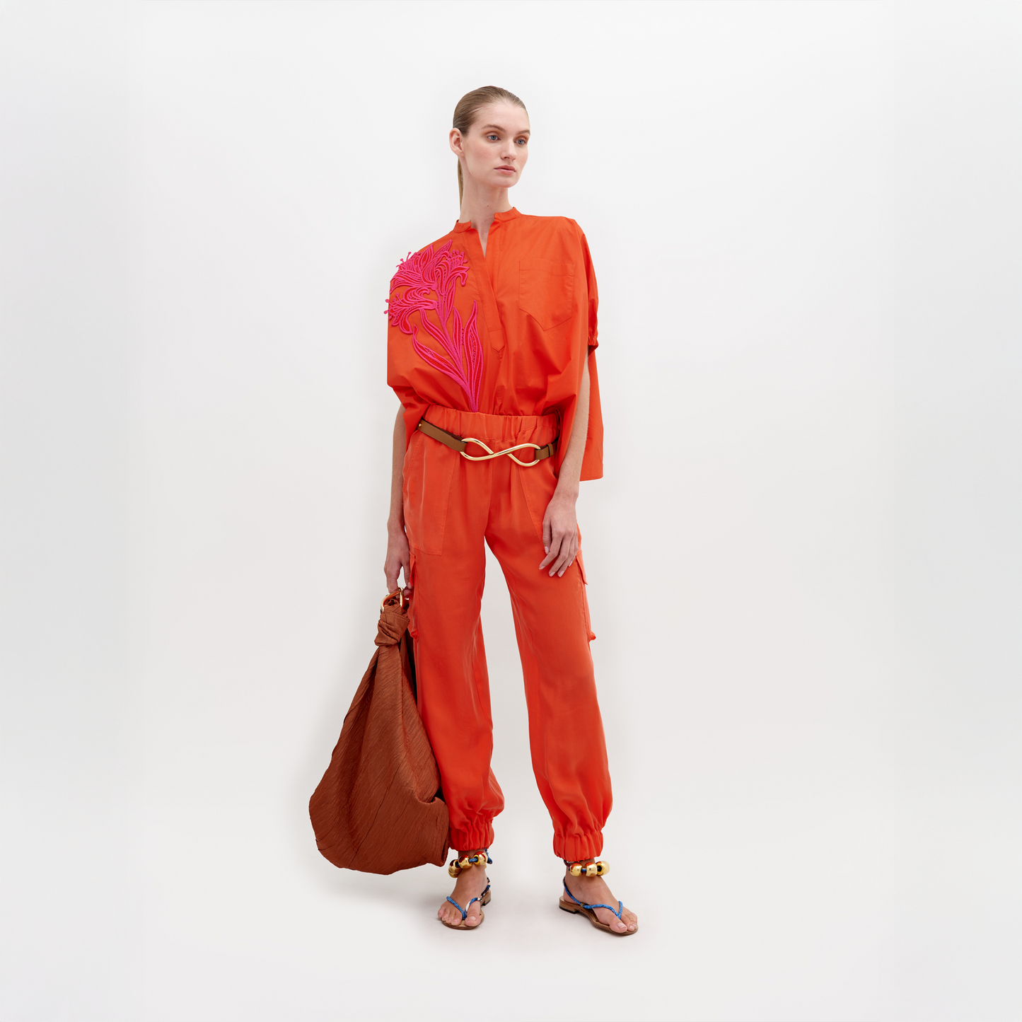 Una persona está parada sobre un fondo blanco vistiendo Pantalón Jess, pantalones holgados de color naranja brillante con cintura con cinturón, combinados con pulseras de tobillo doradas, sandalias azules y sosteniendo un bolso marrón grande.