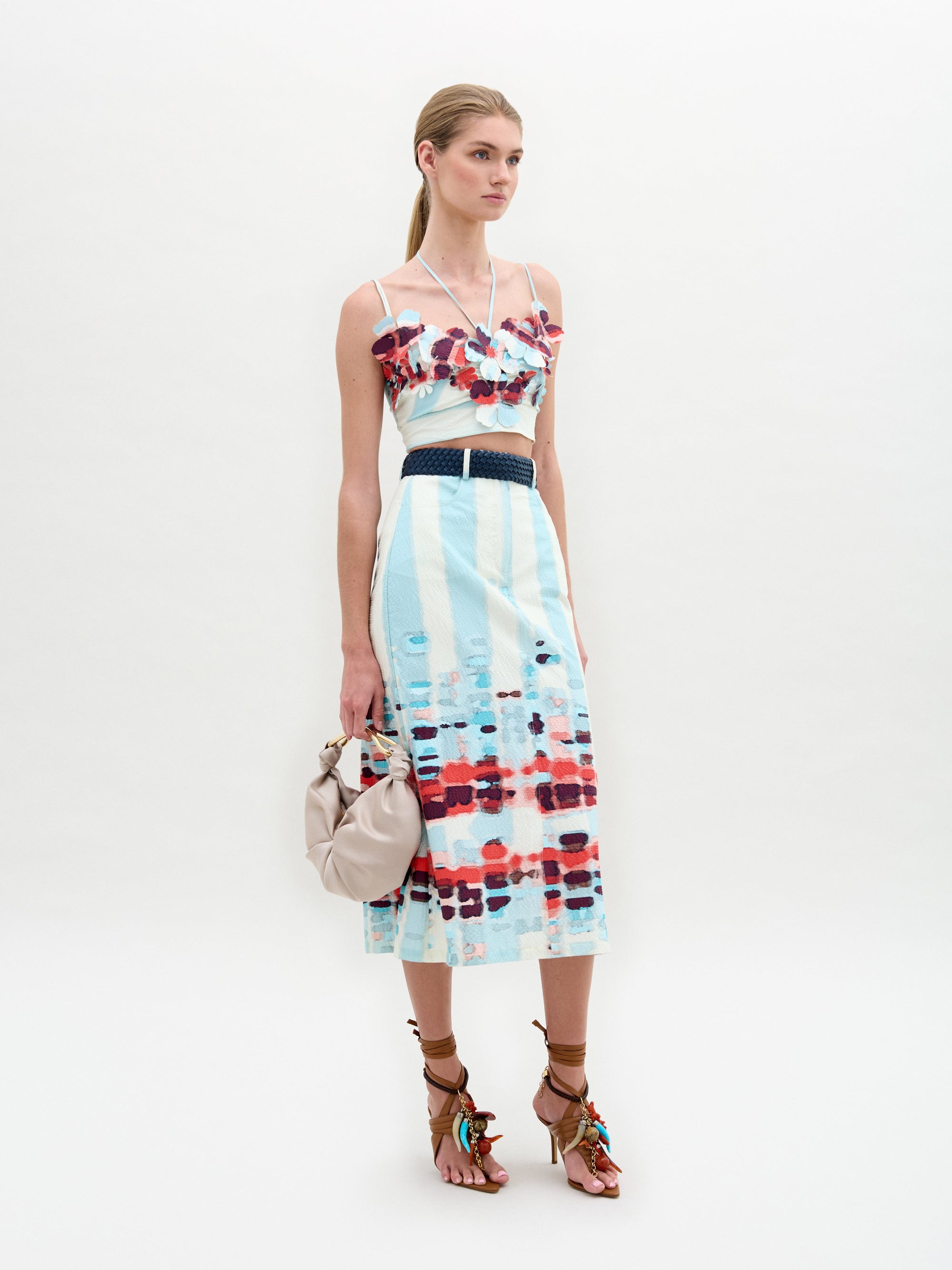 Una mujer está parada sobre un fondo liso con el vestido sin mangas Falda Layan, con estampados en azul, blanco y rojo confeccionado en tela italiana. Sostiene un bolso beige y usa sandalias marrones con borlas de colores. Lleva el cabello recogido.