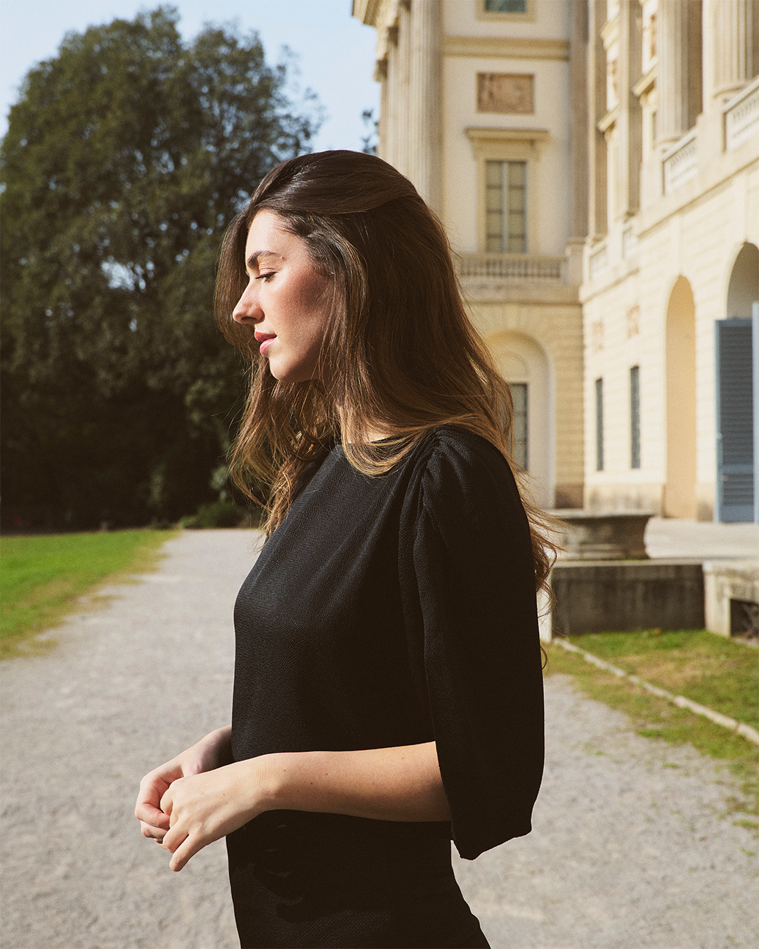 Una mujer de pelo largo, vestida con un vestido negro, se encuentra de pie en un camino de grava junto a un edificio histórico con ventanas arqueadas. Al fondo hay árboles y césped, y la luz del sol proyecta un cálido resplandor.