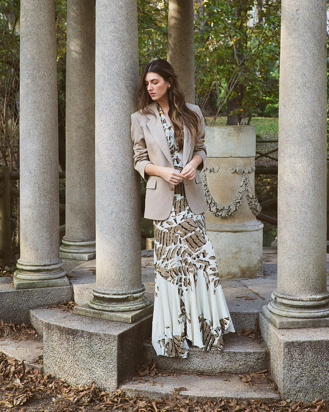 Una mujer con un vestido estampado y una chaqueta beige se encuentra de pie sobre unos escalones de piedra entre columnas. Está al aire libre, rodeada de árboles y hojas caídas.