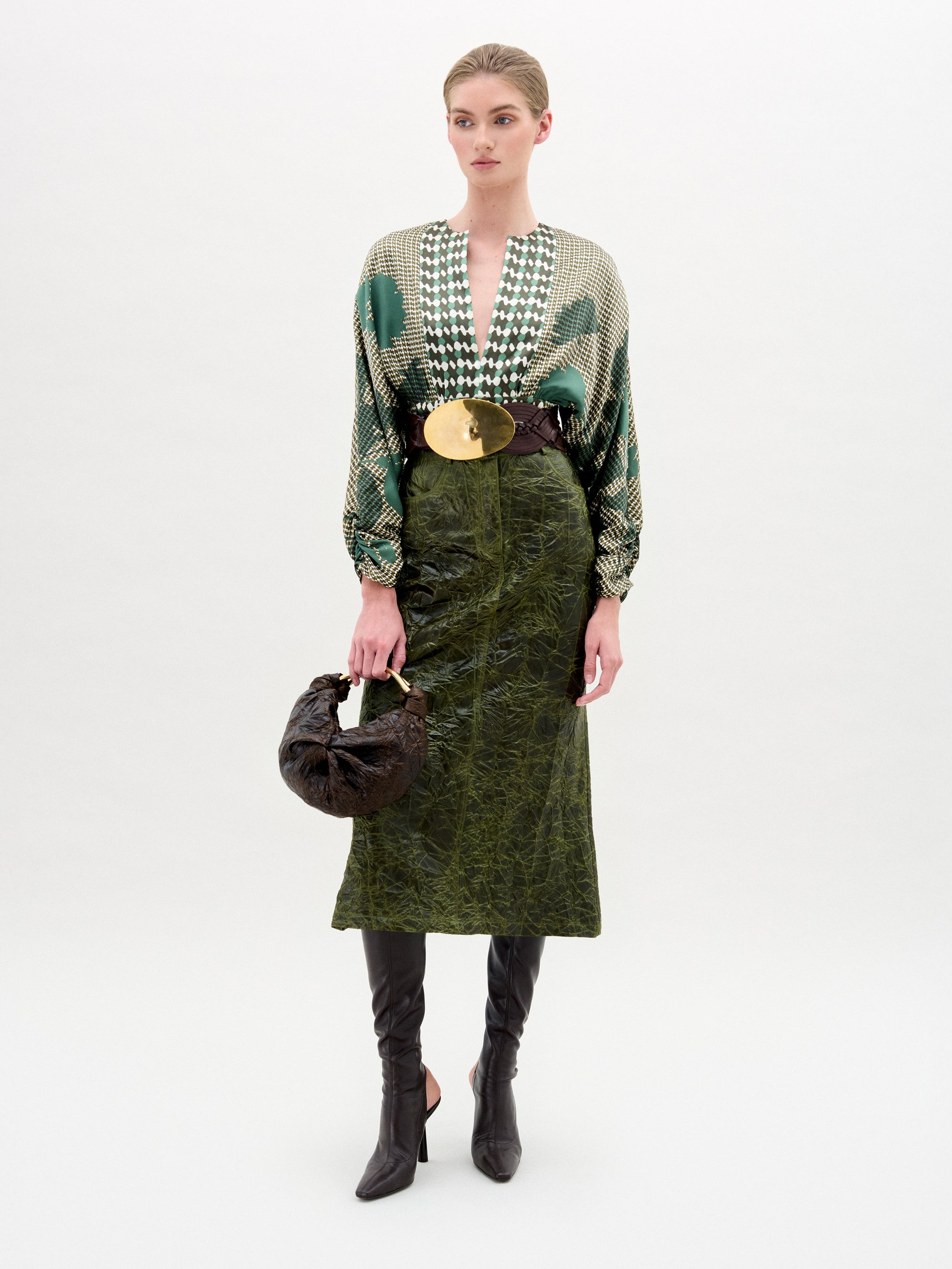 Una persona de pie sobre un fondo liso lleva una blusa verde estampada, un cinturón ancho con una gran hebilla dorada, la falda midi de tela italiana (Layan Skirt), botas negras hasta la rodilla y un pequeño bolso oscuro. Hecho en Colombia.