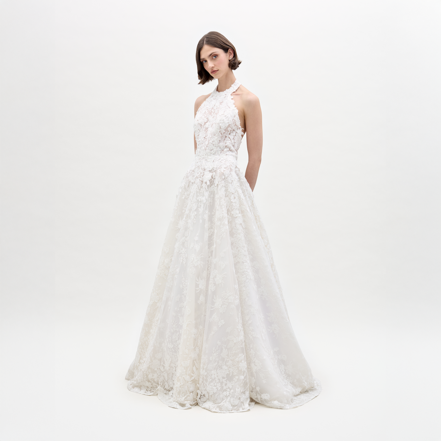 Una mujer se encuentra en un estudio con el Vestido Leia: un vestido de novia blanco, largo hasta el suelo y sin mangas, con encaje translúcido y un elegante escote halter. Lleva el pelo corto y liso, las manos a la espalda y el fondo es blanco liso.