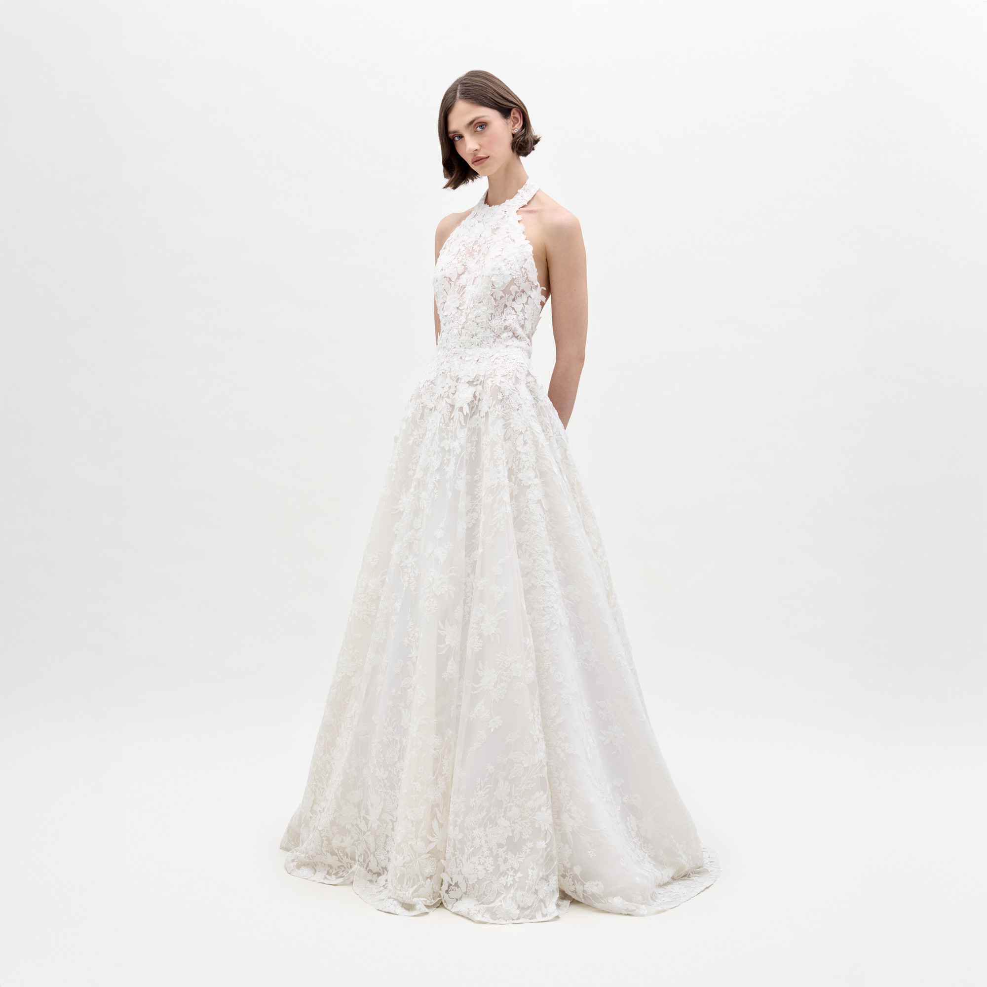 Una mujer se encuentra en un estudio con el Vestido Leia: un vestido de novia blanco, largo hasta el suelo y sin mangas, con encaje translúcido y un elegante escote halter. Lleva el pelo corto y liso, las manos a la espalda y el fondo es blanco liso.