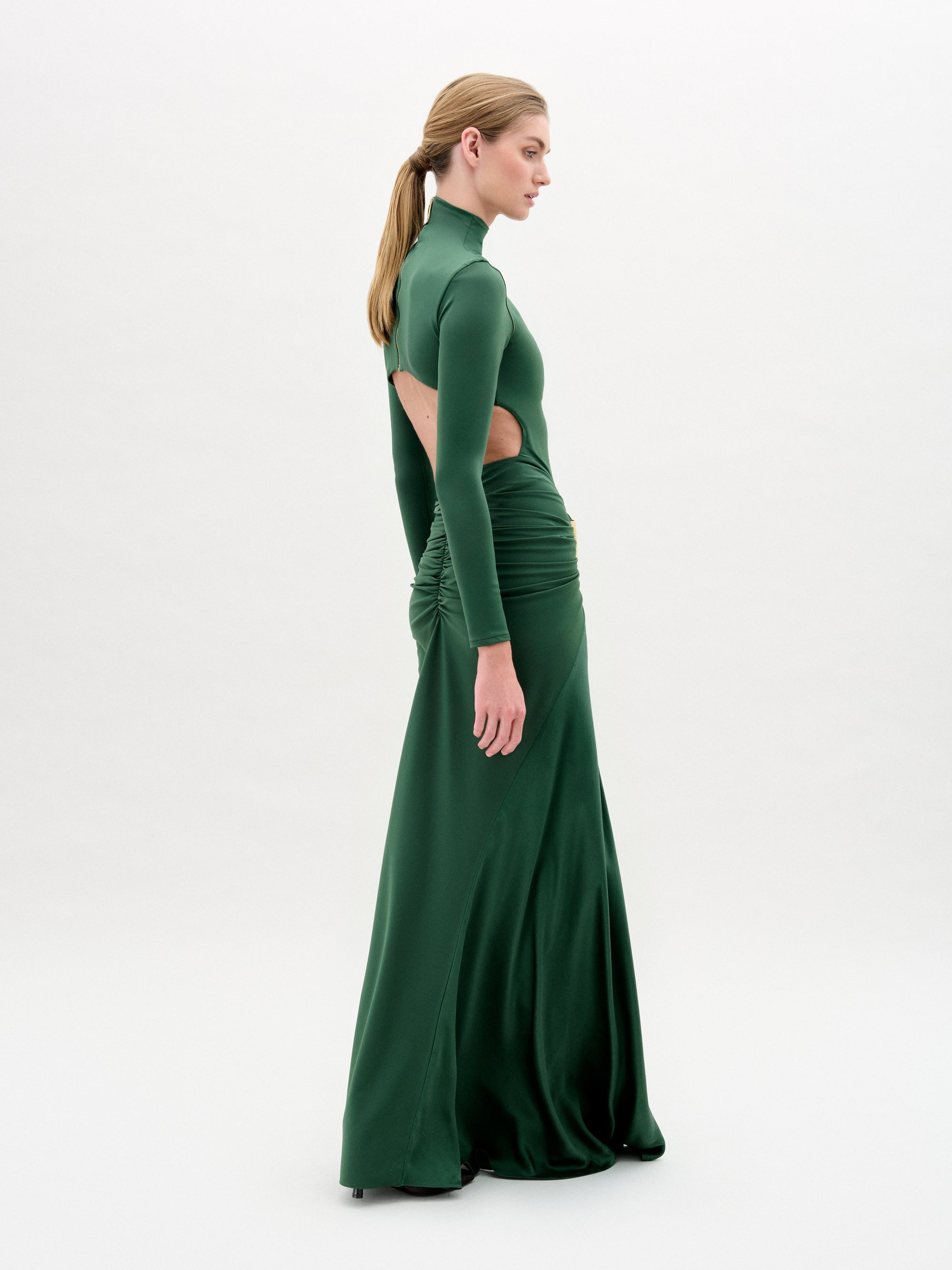 Una persona de perfil luce el Lena Dress, un maxi vestido de talle ajustado en verde oscuro con mangas largas, cuello alto, aberturas laterales y fruncido en la cintura, hecho en Colombia, sobre fondo blanco.