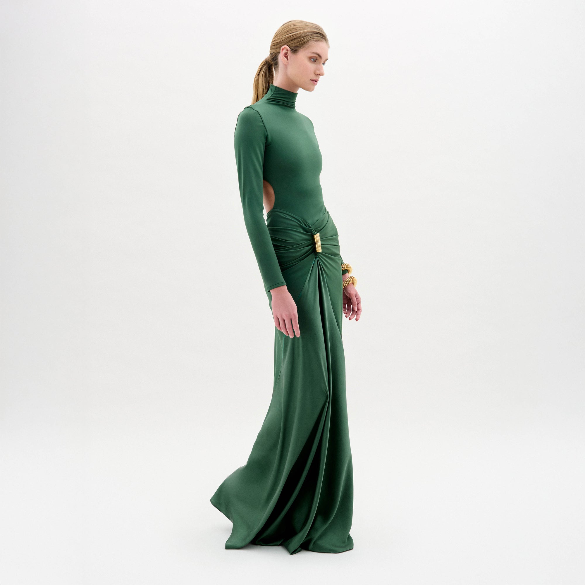 Una mujer de piel clara y pelo rubio recogido en una coleta hace de modelo del vestido Lena, un maxivestido verde con cintura entallada, manga larga, aberturas laterales y detalles fruncidos. Posa de perfil sobre un fondo blanco liso. Fabricado en Colombia.
