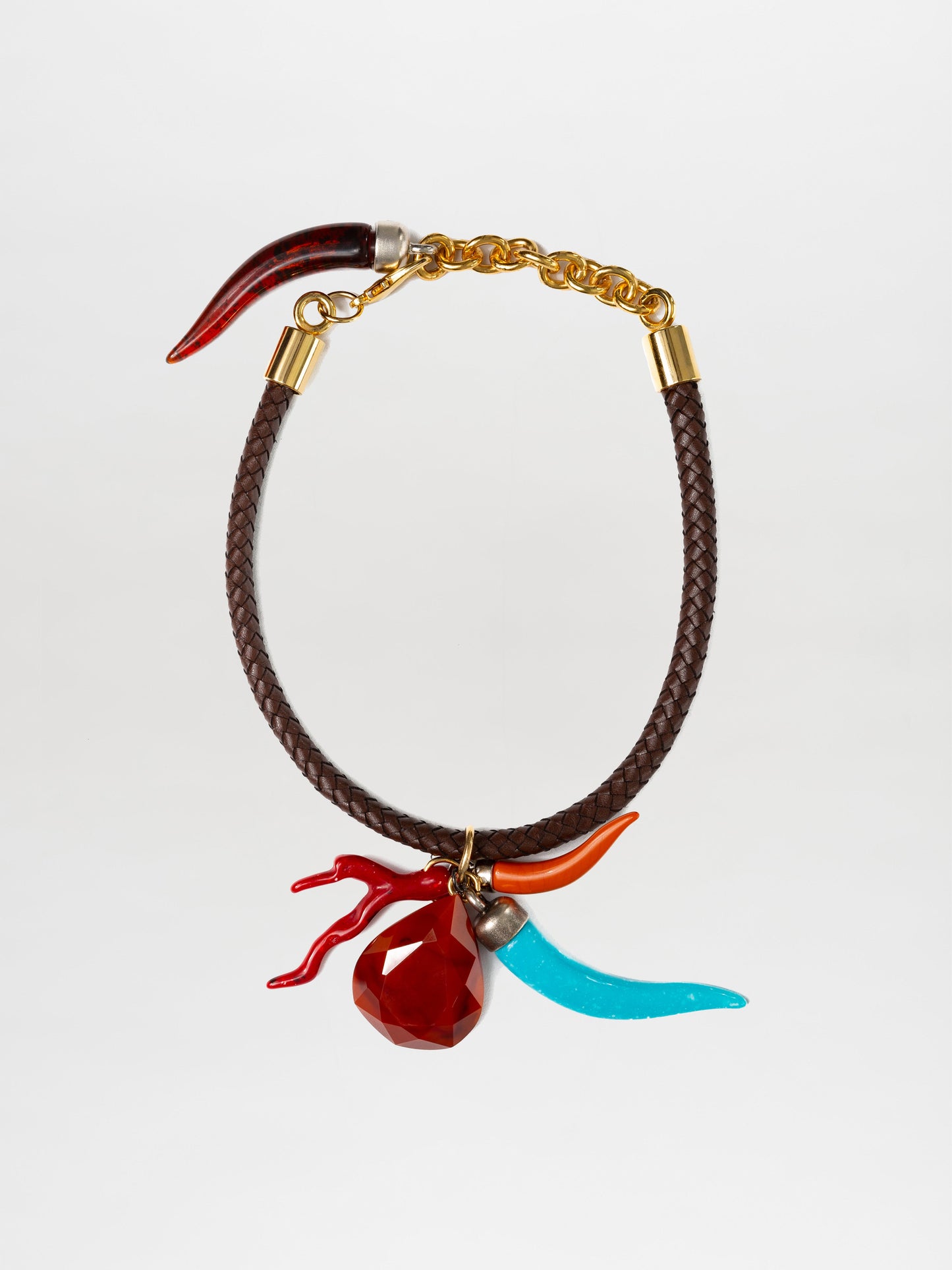 El collar Leslie presenta una banda marrón trenzada con detalles metálicos, un cierre de cadena dorada y tres dijes: una joya roja, un cuerno rojo y un cuerno azul. Los adornos multicolores están unidos cerca del cierre con anillos dorados.