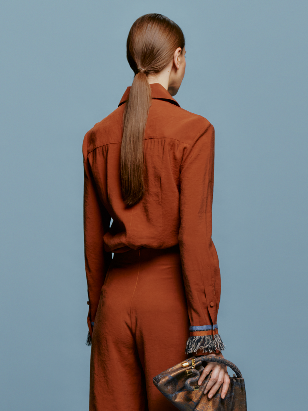 A person stands against a blue background wearing the Liv Blouse Cacao, a rust-colored outfit with fringe details on the collar, front, cuffs, and waistband, highlighting the latest trend expected in the September 2nd shipment. They're holding a small gray handbag.