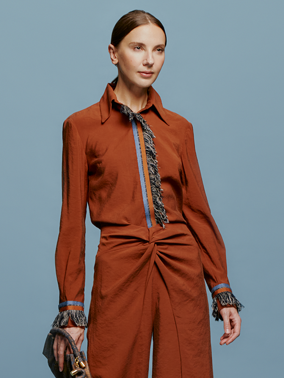 A person stands against a blue background wearing the Liv Blouse Cacao, a rust-colored outfit with fringe details on the collar, front, cuffs, and waistband, highlighting the latest trend expected in the September 2nd shipment. They're holding a small gray handbag.