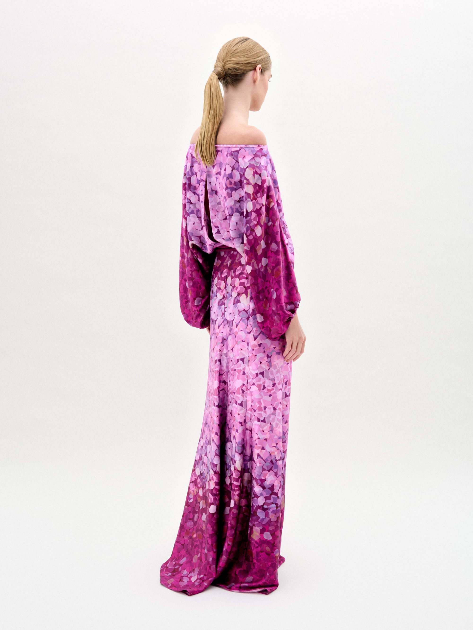 Una persona de piel clara y pelo rubio recogido está de espaldas, con el vestido Lorella -un maxi confeccionado en Colombia con estampados rosas y morados, mangas sueltas y hombros al descubierto- sobre un fondo blanco liso.