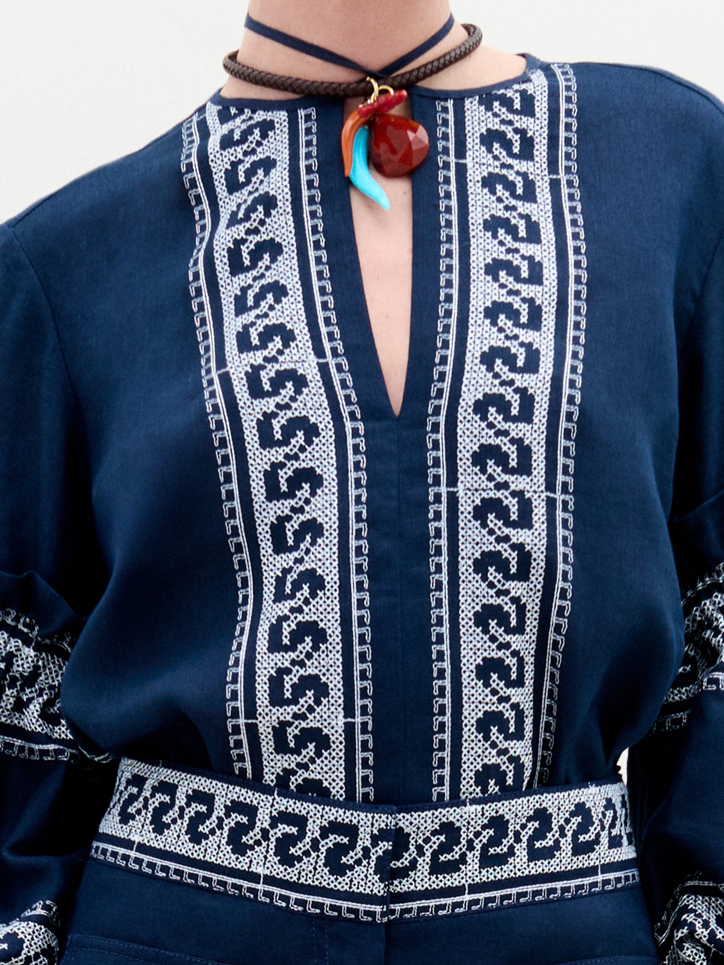 Una persona viste la Blusa Lucaya, una blusa de lino azul marino con intrincados bordados blancos en la parte delantera y las mangas. Su escote en forma de V resalta hermosamente un collar rojo con colgante. Esta exquisita pieza está elaborada por expertos en Colombia.