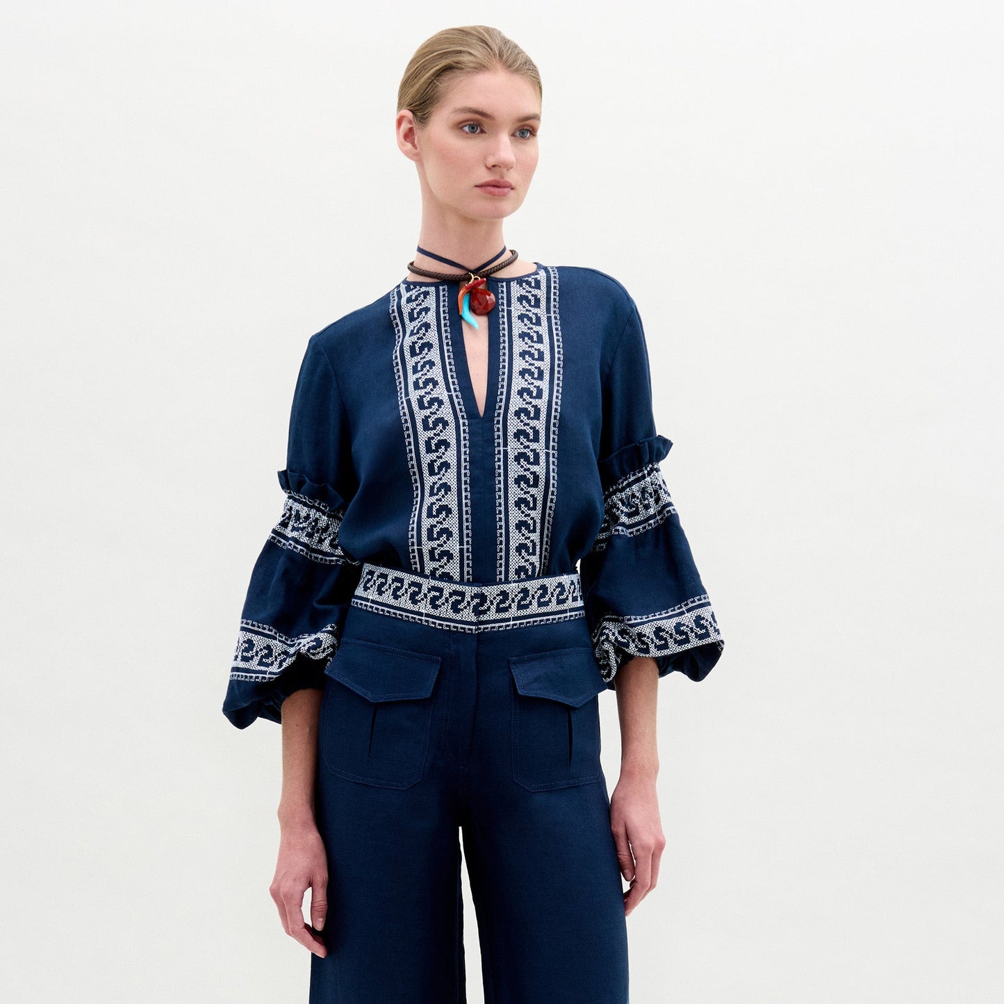 Con la blusa Lucaya de lino azul marino con bordados blancos y mangas voluminosas, combinada con unos pantalones de talle alto a juego, una persona se alza sobre un fondo claro liso.