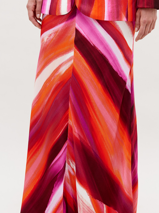 A person stands against a plain background, wearing a vibrant suit in red, pink, and orange streaks, paired with the Laurina Skirt Magenta Abstract Waves and bold gold necklace. This stunning ensemble will be available for pre-order soon and is set to ship by date 2024.