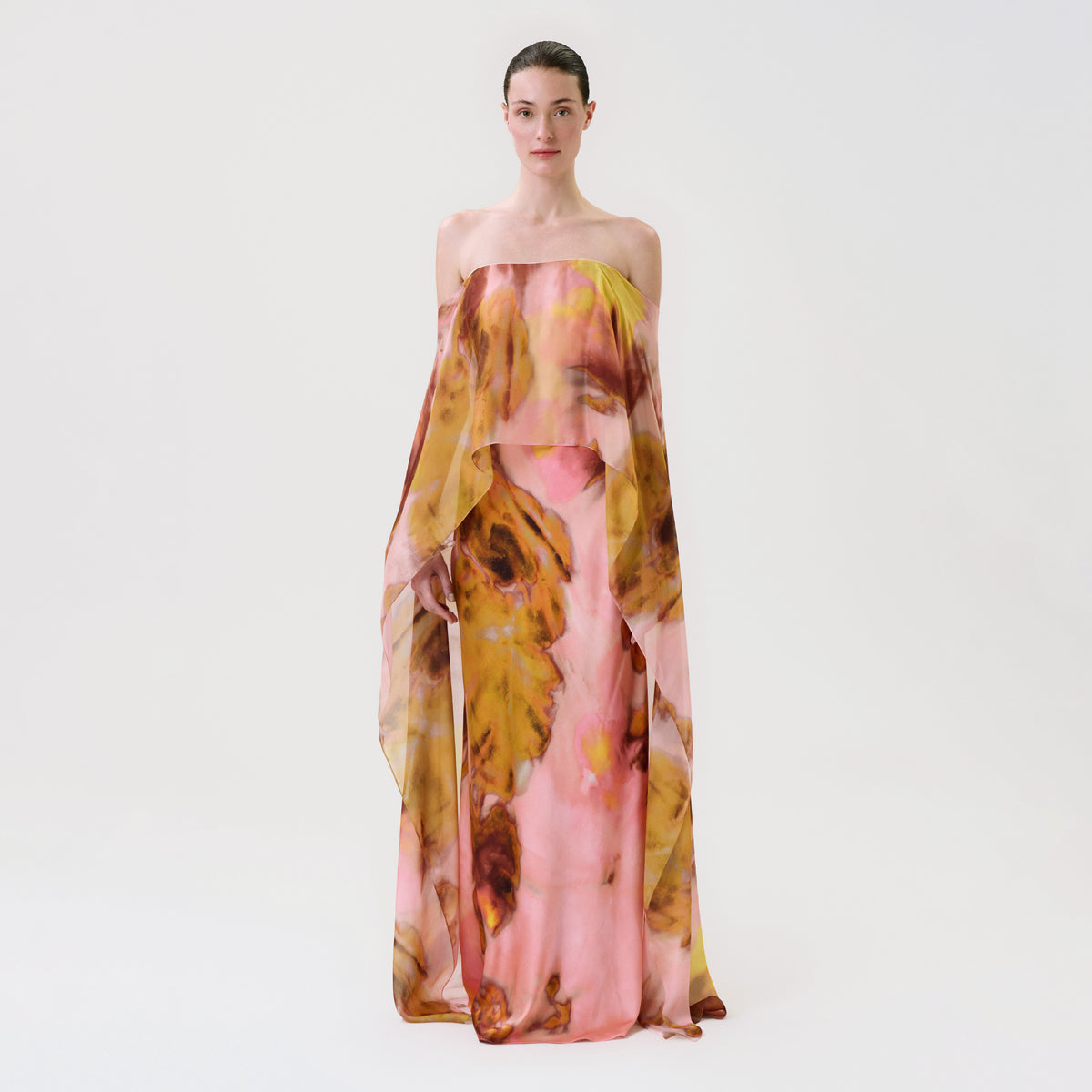 A person stands against a plain background wearing the strapless, flowing Lovane Dress, featuring a pink, yellow, and brown abstract pattern.