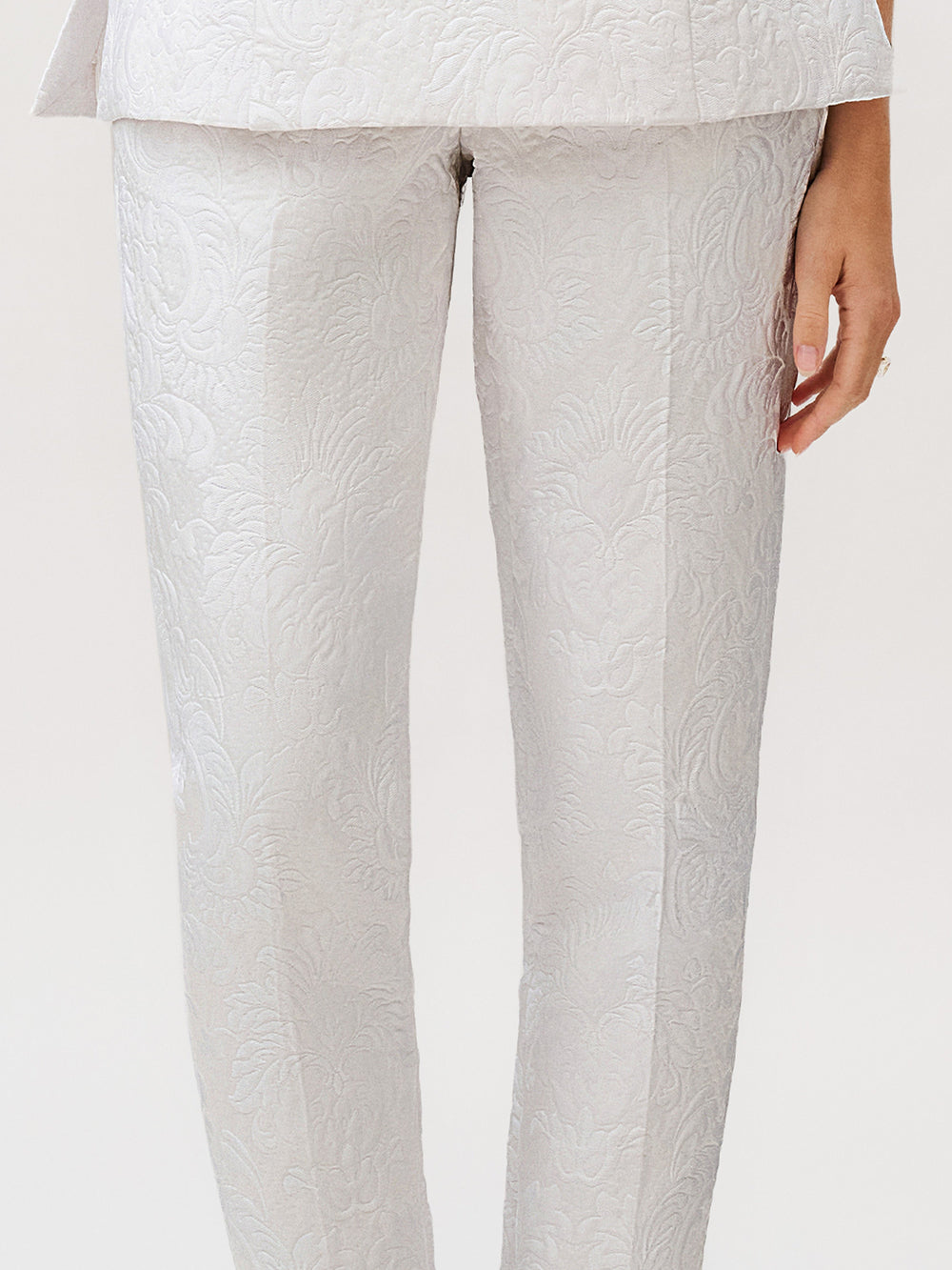 A person wearing the Luca Pant in light gray silk cotton brocade with a textured floral pattern, paired with a matching jacket—an elegant ready-to-wear ensemble made in Italy, with one hand visible by their side.