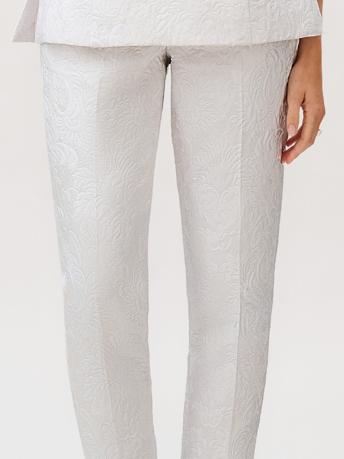 A person wearing the Luca Pant in light gray silk cotton brocade with a textured floral pattern, paired with a matching jacket—an elegant ready-to-wear ensemble made in Italy, with one hand visible by their side.