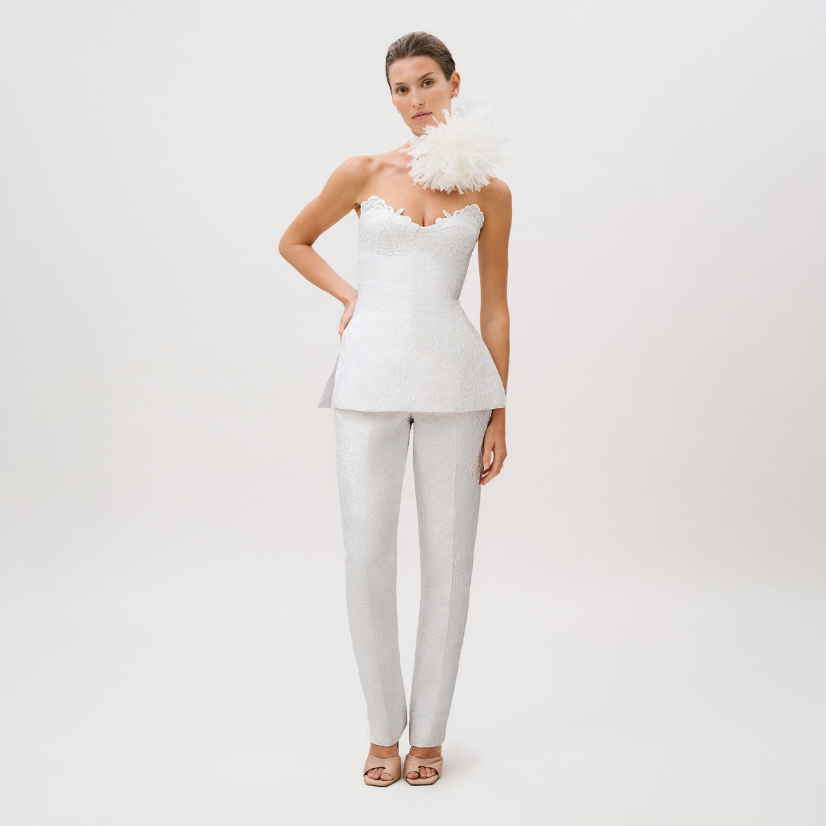 A woman models the Luca Pant—a pair of matching fitted trousers—styled with a textured white strapless peplum top featuring a large white floral accent at the shoulder, crafted from exquisite silk cotton brocade, and finished with open-toed heels. The ready-to-wear ensemble is elegantly posed against a plain light background.