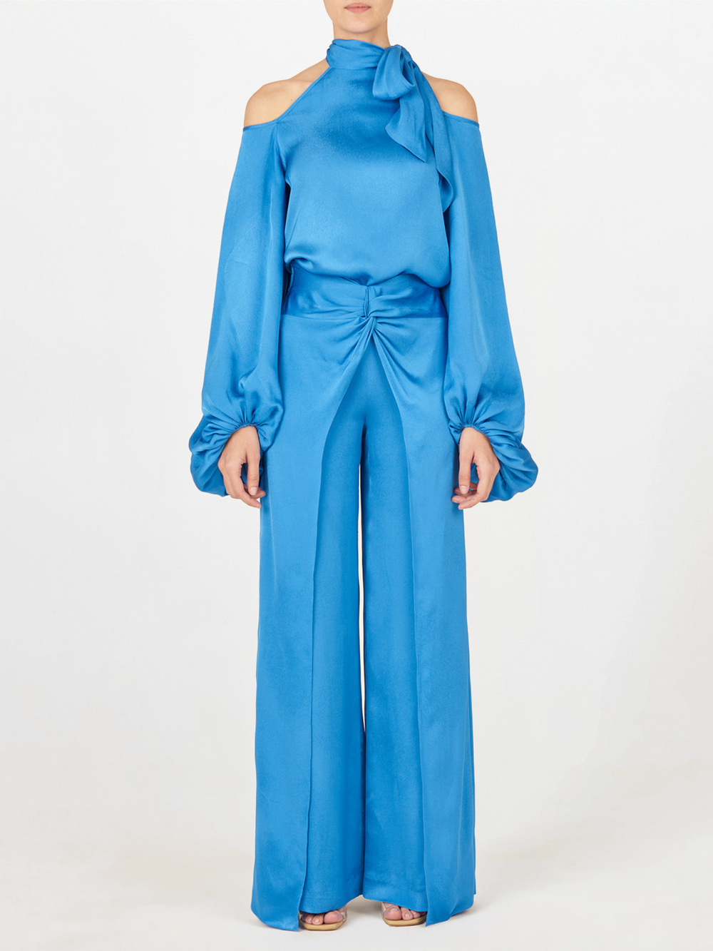 Vestida con la Blusa Ludovica Azul con pantalones de pierna ancha, mangas largas abullonadas, recortes en los hombros fríos y escote alto con lazo, una persona se alza sobre un fondo blanco liso.