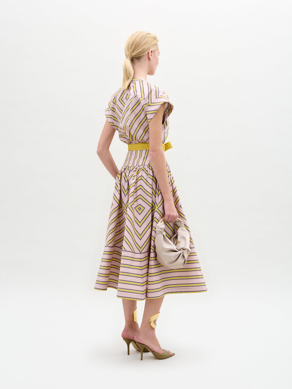 Una mujer con el pelo rubio recogido en una coleta baja está de espaldas, lleva el vestido Macarena con motivos geométricos, tacones altos y sostiene un bolso de mano recogido de color claro. El fondo es liso y neutro.