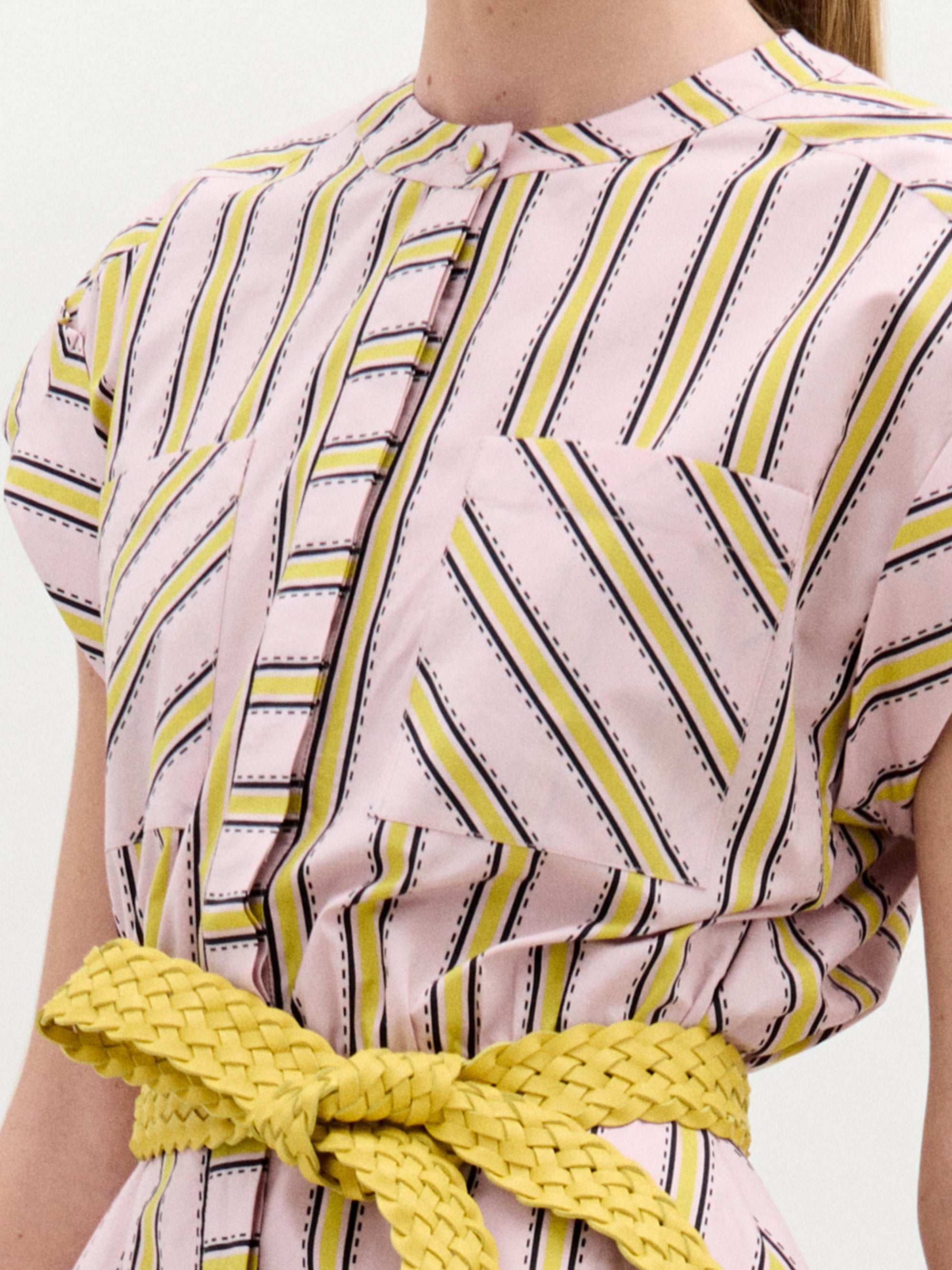 Una persona lleva el Vestido Macarena, un vestido rosa claro de manga corta con rayas diagonales amarillas, negras y blancas, un bolsillo delantero y un cinturón amarillo trenzado en la cintura. La imagen está recortada por los hombros y la cintura.