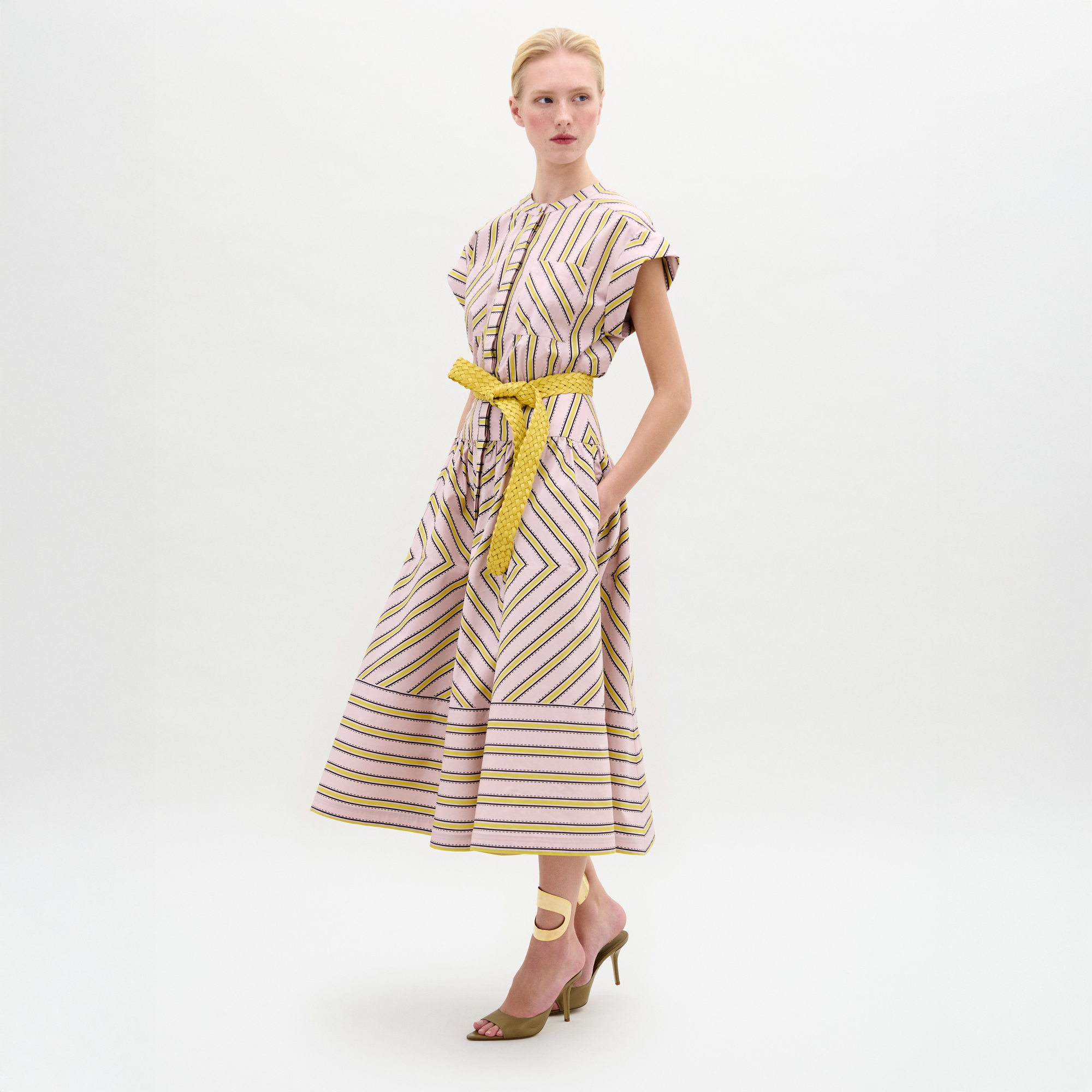 Una mujer posa sobre fondo blanco con el vestido Macarena, de largo medio y manga corta, con rayas geométricas y cinturón amarillo. Lleva una mano en el bolsillo y completa el look con sandalias de tacón.