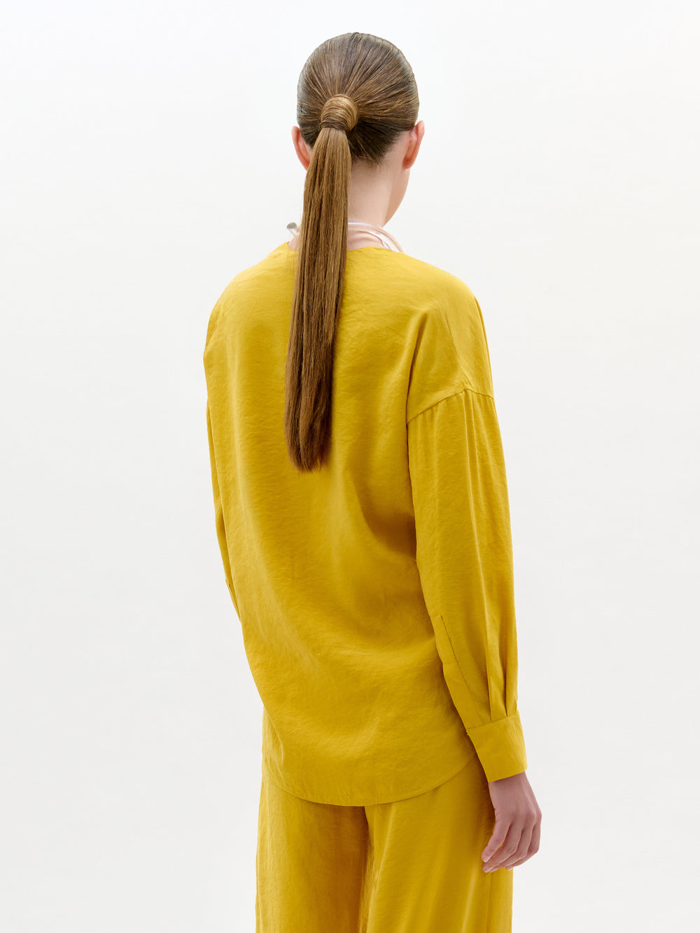 Una persona con el pelo largo y castaño recogido en una coleta baja aparece de espaldas, vistiendo la blusa de Mérida en amarillo con pantalones a juego sobre un fondo claro liso.