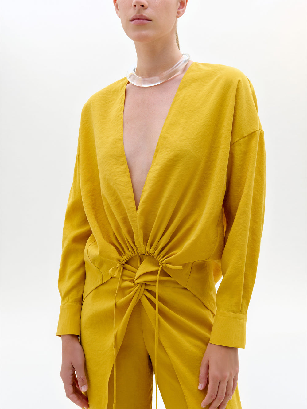 Sobre un fondo blanco liso, una persona luce la blusa Mérida, un top holgado de color amarillo mostaza con mangas largas, escote profundo en V y cintura con cordón, combinado con un grueso collar translúcido.