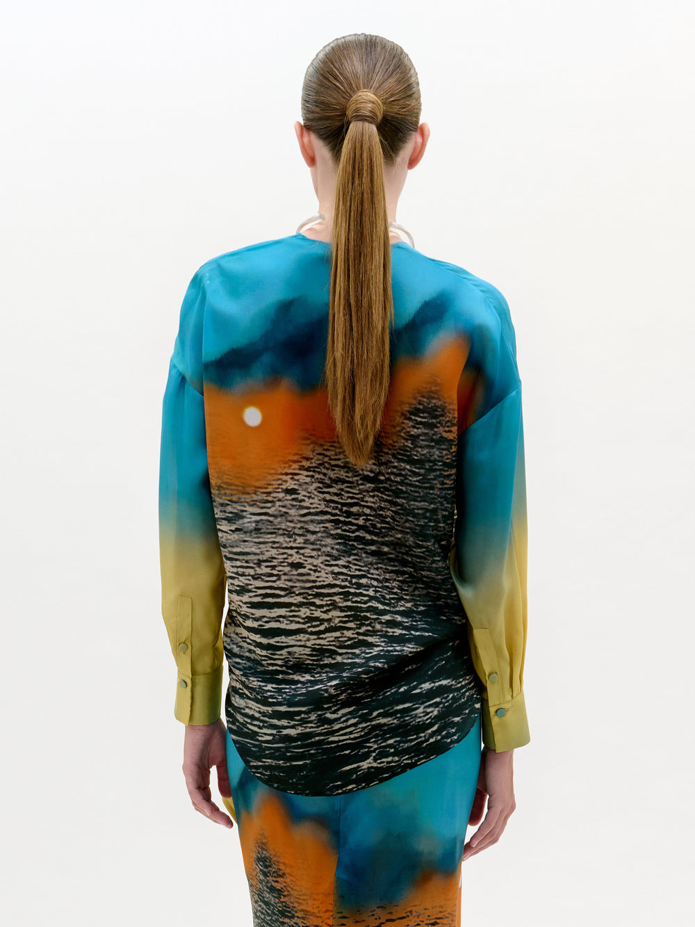 Una persona con el pelo largo y castaño recogido en una coleta baja aparece de espaldas, vistiendo la blusa Mérida, una blusa de manga larga con un estampado de atardeceres oceánicos en tonos azules, naranjas y amarillos. El fondo es liso y de color claro.