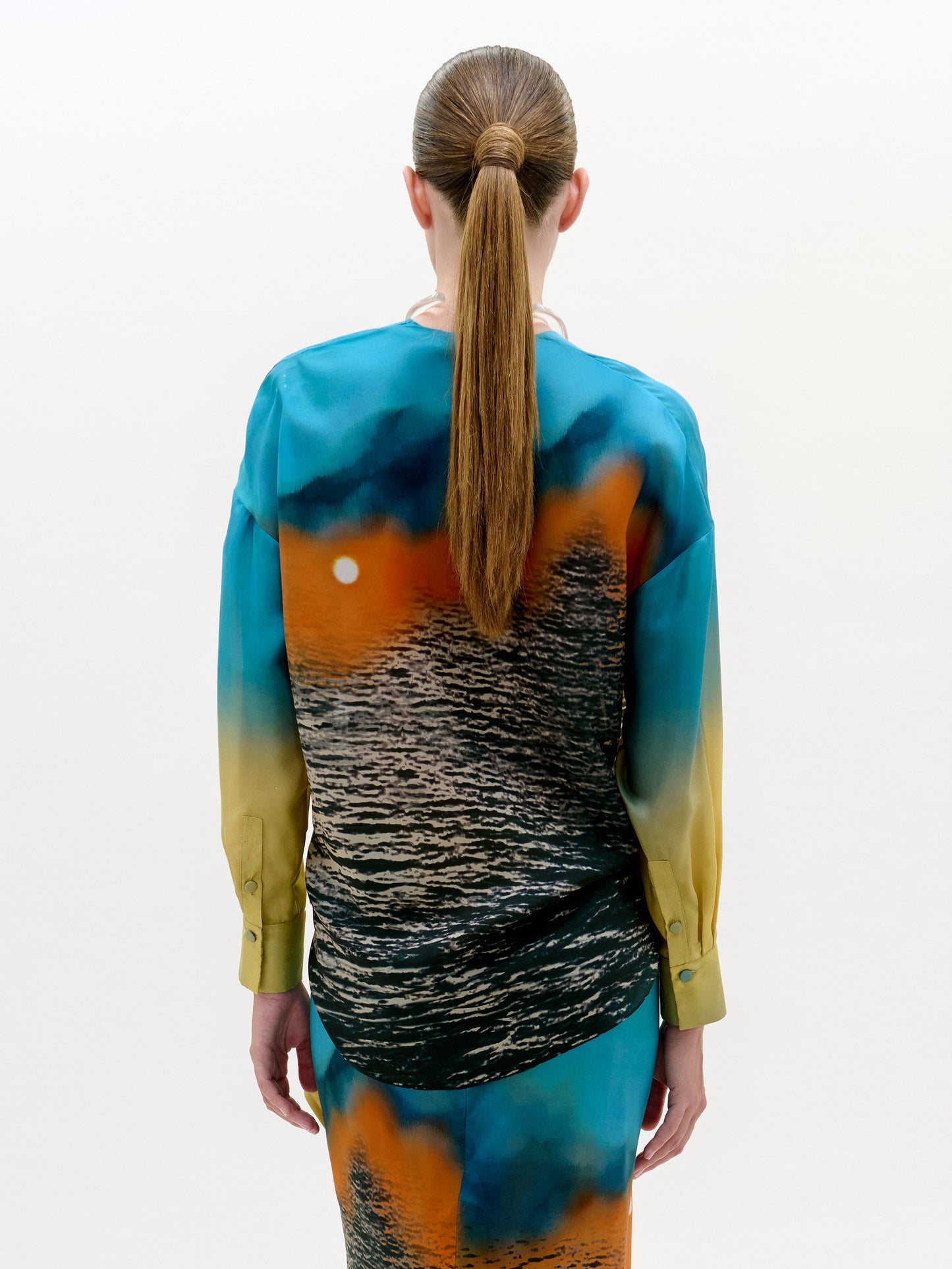 Una persona con el pelo largo y castaño recogido en una coleta baja aparece de espaldas, vistiendo la blusa Mérida, una blusa de manga larga con un estampado de atardeceres oceánicos en tonos azules, naranjas y amarillos. El fondo es liso y de color claro.