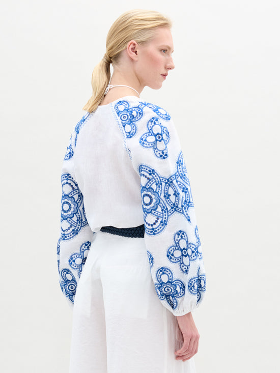 La mujer, que lleva la Blusa Molveno, una blusa blanca con estampados florales azules y un elegante escote en V, combinada con pantalones blancos, aparece de perfil sobre un fondo liso. Lleva el pelo recogido en una cola de caballo.