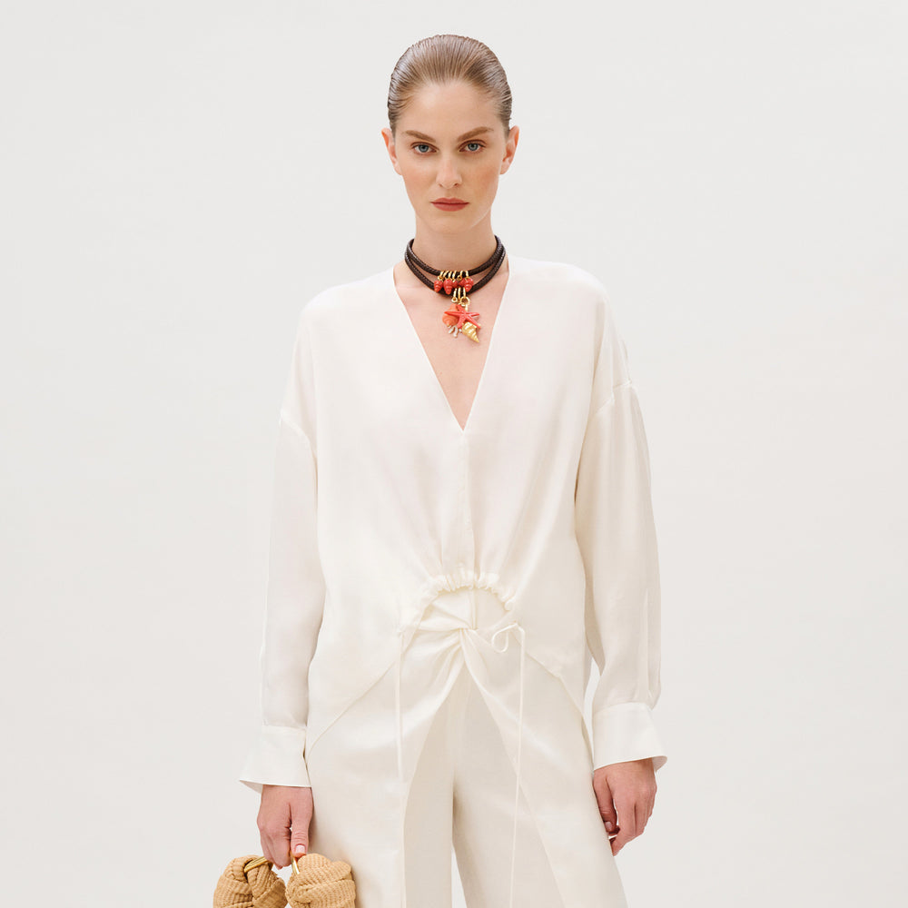 A person wearing a white, long-sleeved Merida Blouse in cupro with a V-neck, a gathered drawstring waist, and a layered beaded necklace featuring a starfish charm.