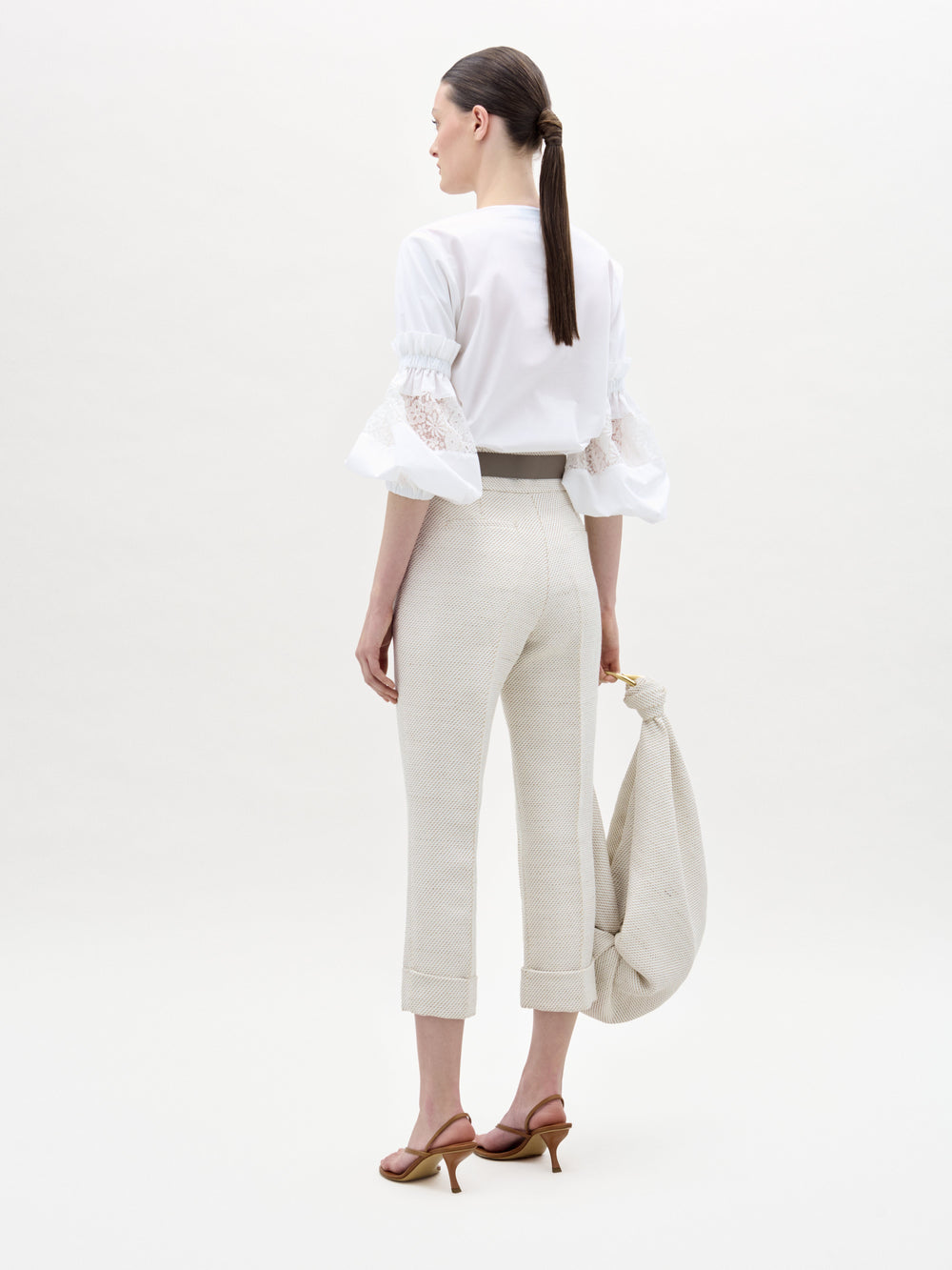 Una mujer, de espaldas, lleva una blusa blanca con mangas de encaje y el pantalón Moad en beige claro, combinado con sandalias de tacón marrones. Lleva un bolso de tela beige y el pelo recogido en una coleta baja.