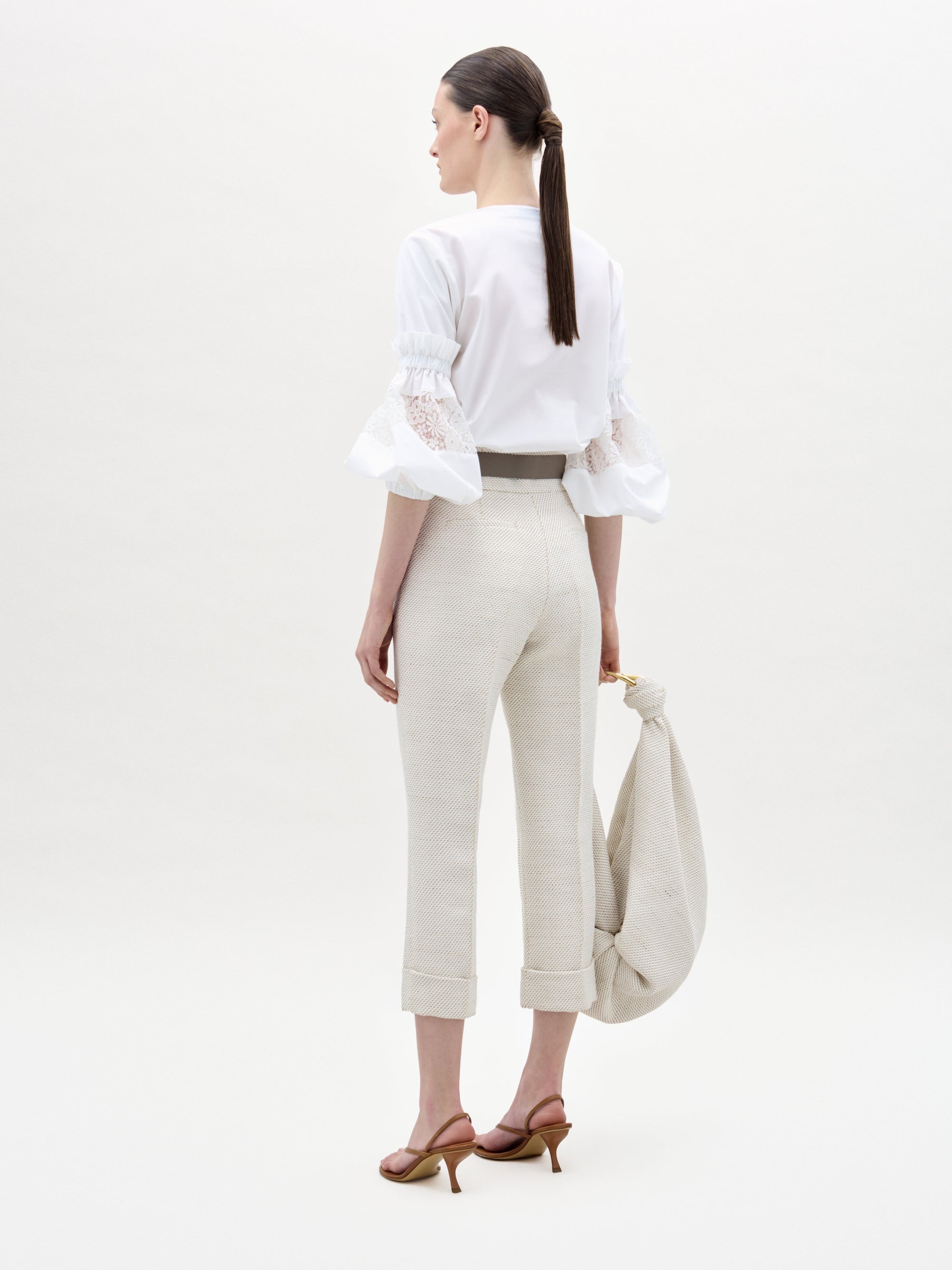 Una mujer, de espaldas, lleva una blusa blanca con mangas de encaje y el pantalón Moad en beige claro, combinado con sandalias de tacón marrones. Lleva un bolso de tela beige y el pelo recogido en una coleta baja.