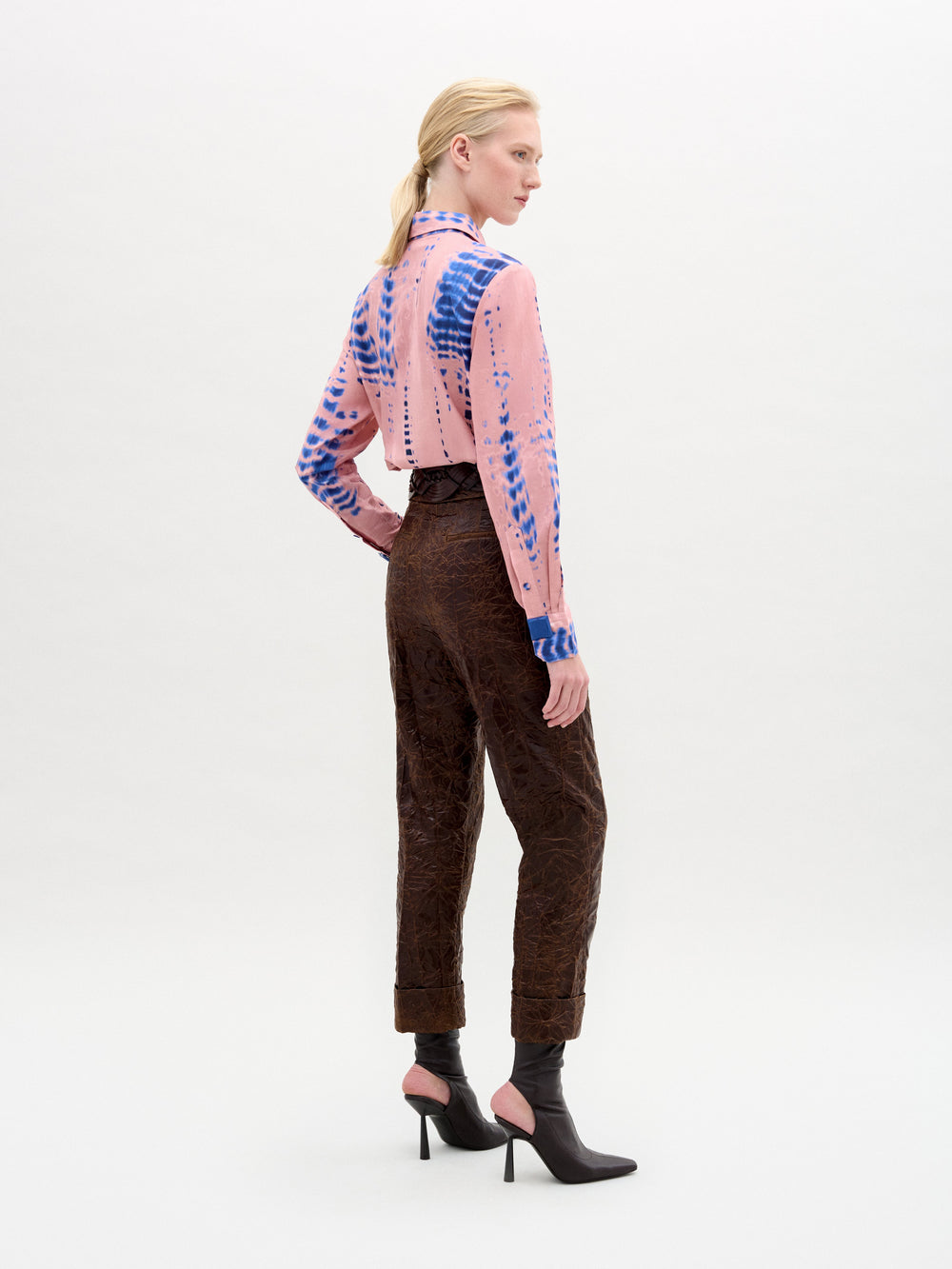 Una mujer, de pie y en ángulo, lleva una camisa rosa estampada, el pantalón Moad Pant -un pantalón de talle alto de tela italiana fabricado en Colombia- y tacones negros con tobilleras rosas, sobre un fondo claro liso.