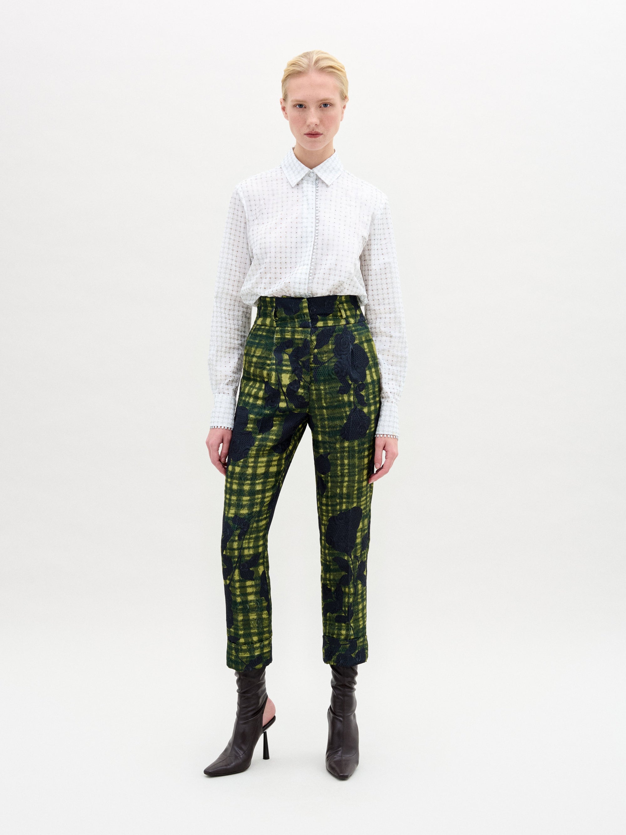 Una persona aparece sobre un fondo blanco liso con la blusa Naroa -100% algodón con diseño de lunares cortado a láser- combinada con unos pantalones de talle alto a cuadros verdes y negros con estampados abstractos, y unos botines de tacón oscuro.