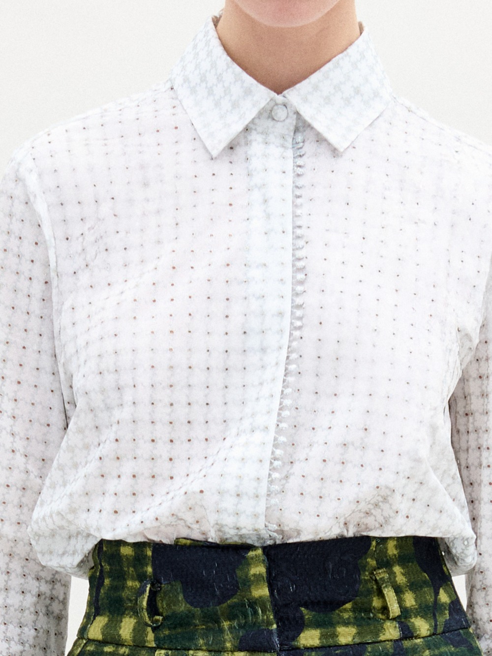 Lleva la blusa Naroa, una blusa blanca de manga larga con un sutil estampado de cuadrícula, metida por dentro de unos pantalones verdes de cintura alta con un diseño abstracto oscuro. El look chic está parcialmente fuera de cuadro.
