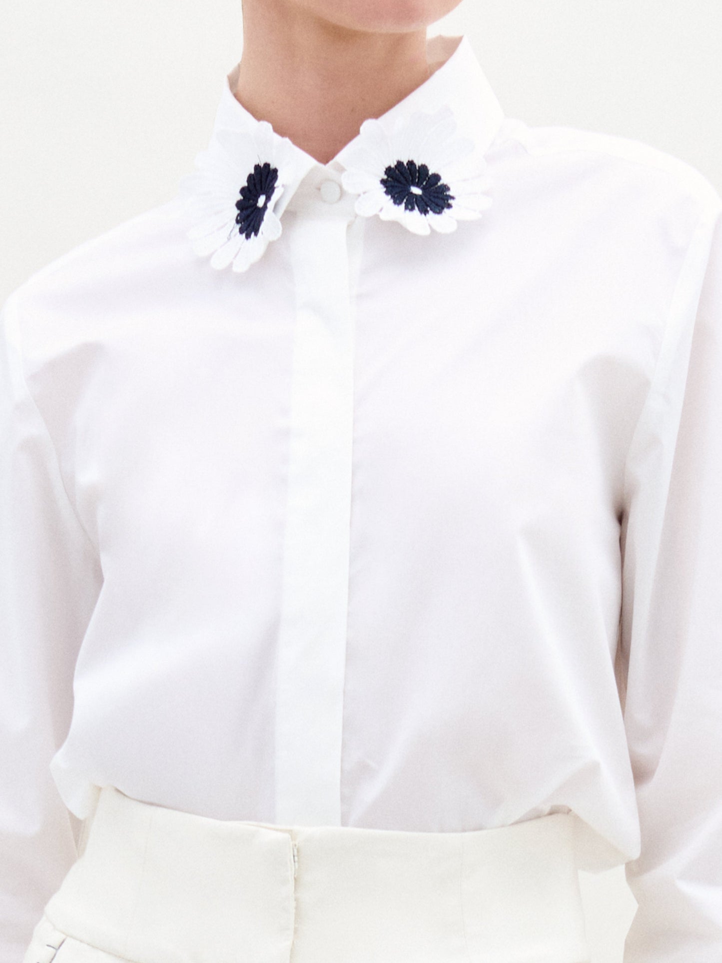 Una persona lleva la Blusa Naroa, una camisa blanca de algodón 100 % con botones y cuello decorativo con intrincados bordados florales en blanco y negro. La imagen resalta los elegantes detalles del diseño en la parte superior del torso.