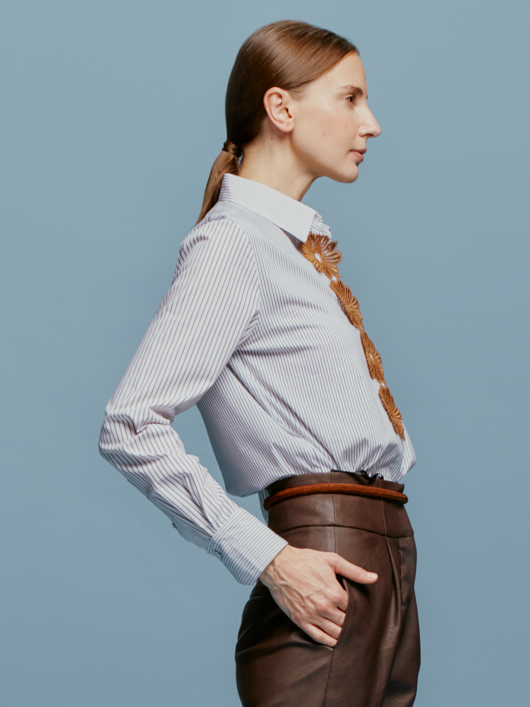 A brown-haired woman with her hair tied back is wearing the Naroa Blouse Multi Brown Stripes, featuring floral embroidery, along with brown pants. She is standing in front of a light blue background, indicating the date as August 19th, 2024.