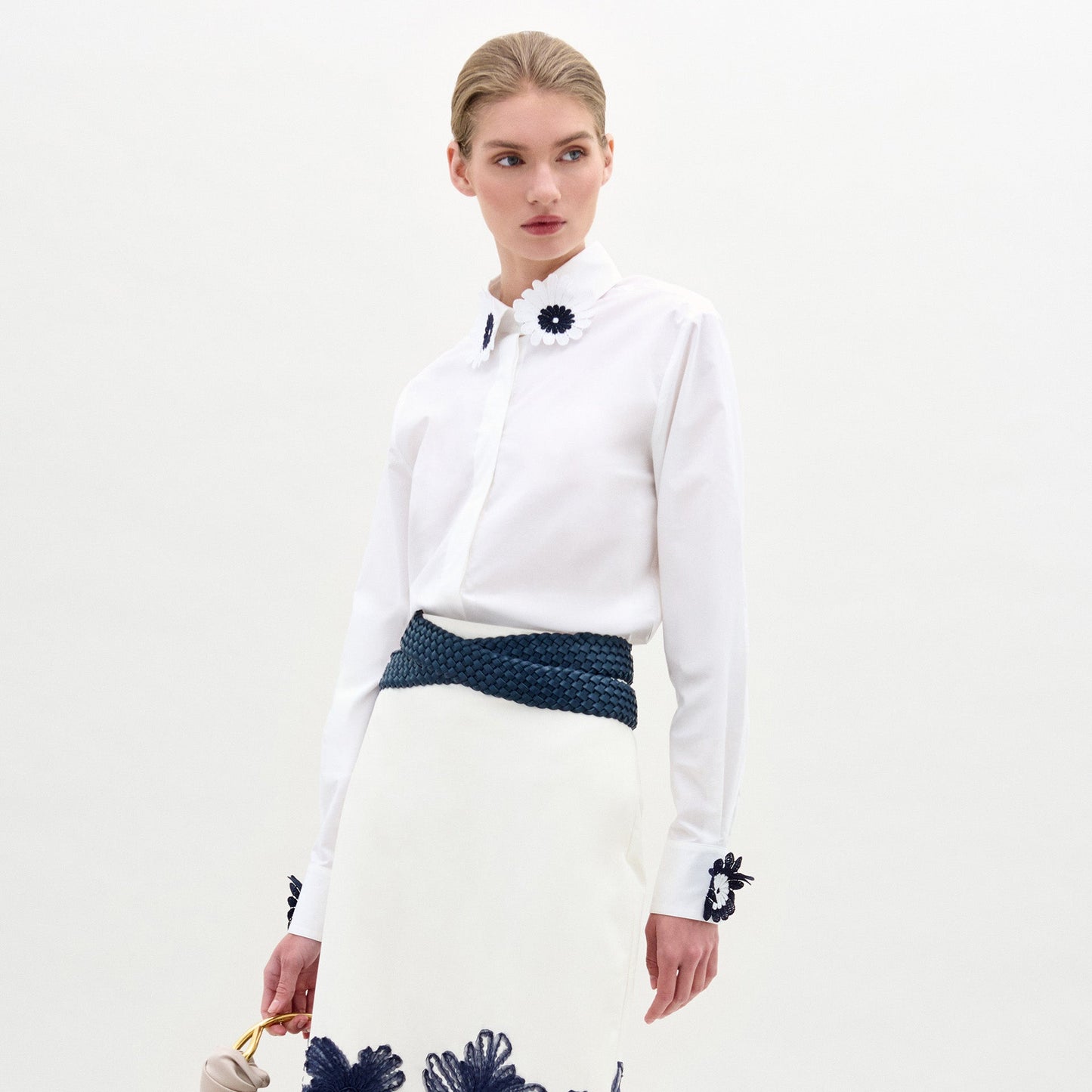 Una persona lleva la blusa Naroa con aplicaciones florales oscuras en el cuello y los puños, combinada con una falda blanca con detalles florales azules en el bajo, un cinturón trenzado azul y un bolso ligero.