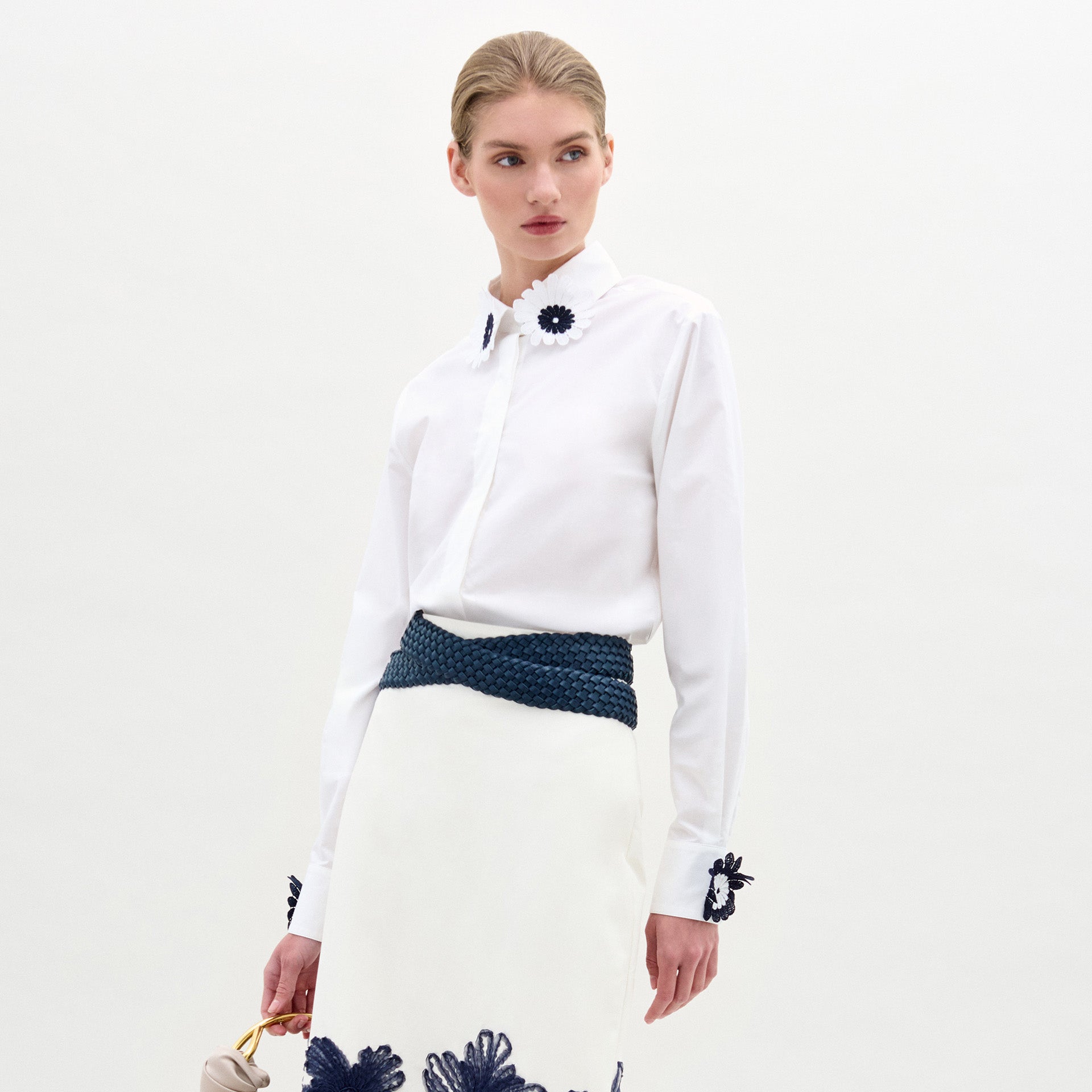 Una persona lleva la blusa Naroa con aplicaciones florales oscuras en el cuello y los puños, combinada con una falda blanca con detalles florales azules en el bajo, un cinturón trenzado azul y un bolso ligero.