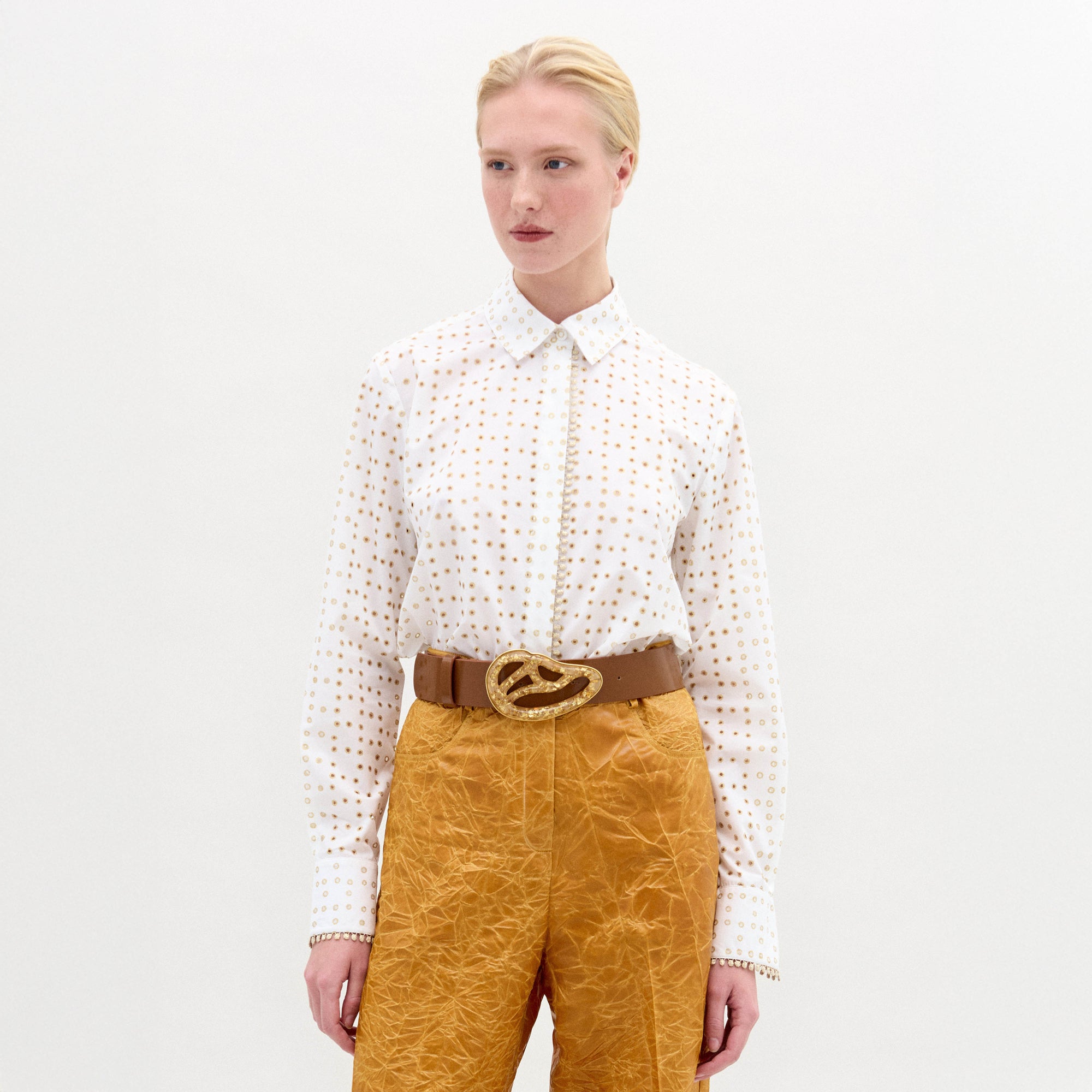 Una persona, sobre un fondo blanco liso, lleva la blusa Naroa con un pequeño estampado de puntos dorados, combinada con unos pantalones dorados arrugados y un cinturón marrón con una gran hebilla dorada decorativa.