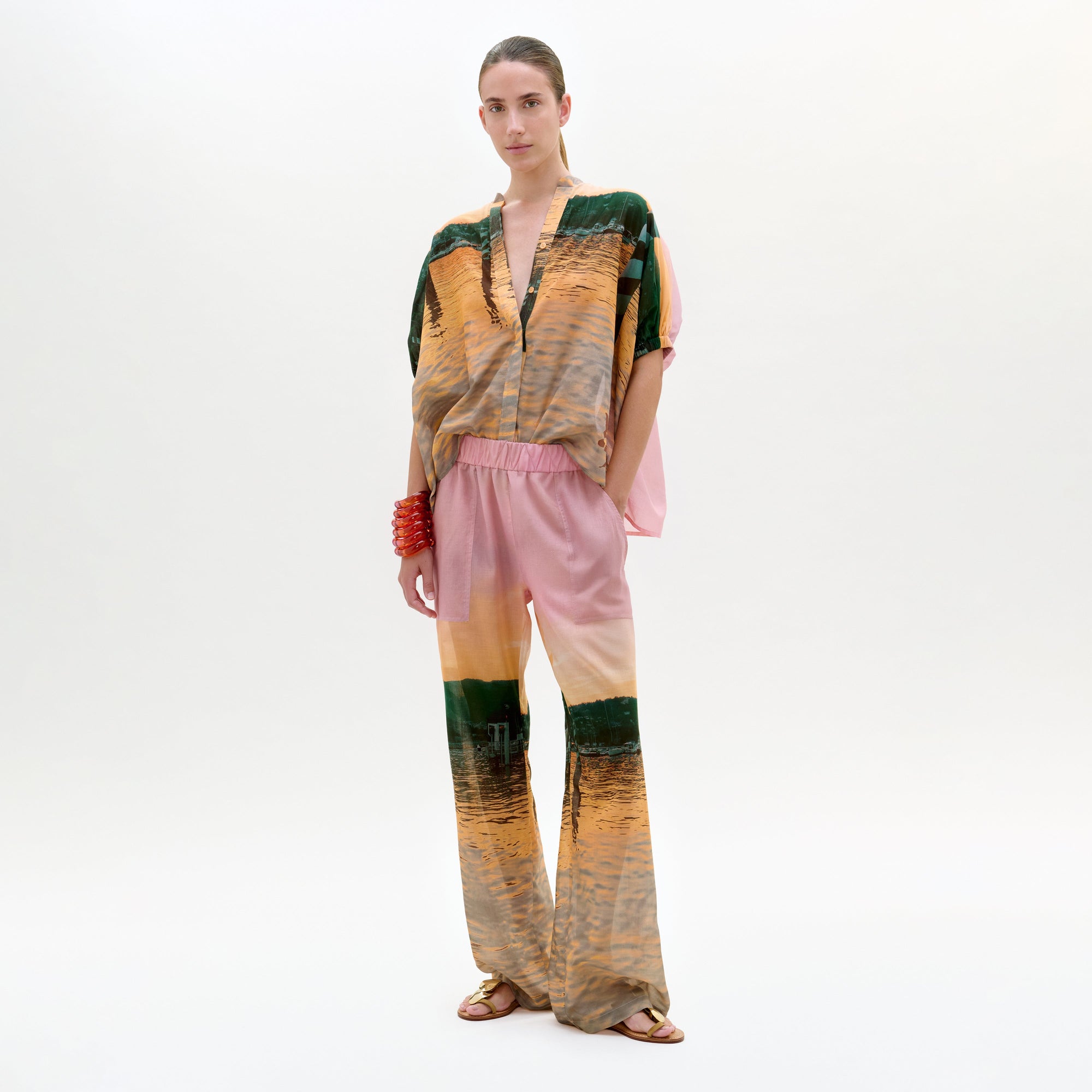 Sobre un fondo liso, una persona lleva el pantalón Nerea, un modelo suelto de cintura alta con un estampado abstracto verde, marrón y rosa. Lo lleva con sandalias, una pulsera roja y el pelo recogido.
