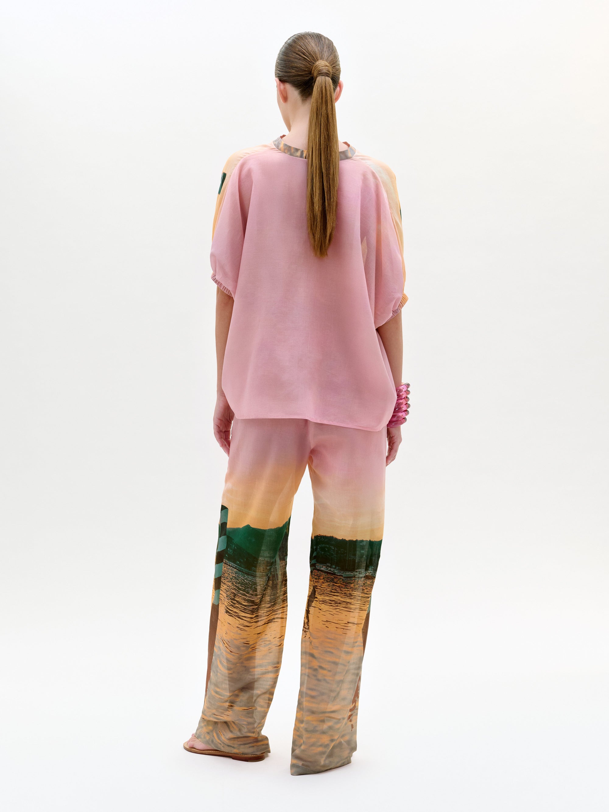 Una persona, con el pelo recogido en una coleta baja y una pulsera gruesa, aparece sobre un fondo blanco liso con un top rosa holgado y el pantalón de cintura alta Nerea Pant, con un estampado de paisajes en tela italiana.