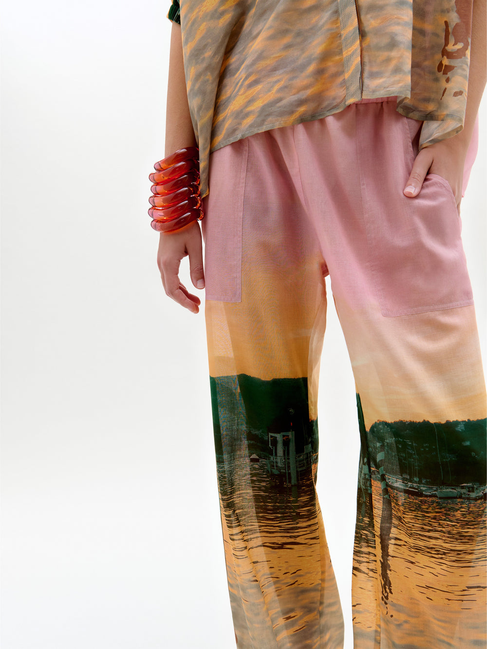 Una persona, con una mano en el bolsillo rosa, lleva el pantalón de talle alto Nerea Pant, hecho en Colombia con un estampado escénico, y un top a juego. Los accesorios son una pulsera roja gruesa sobre un fondo blanco liso.