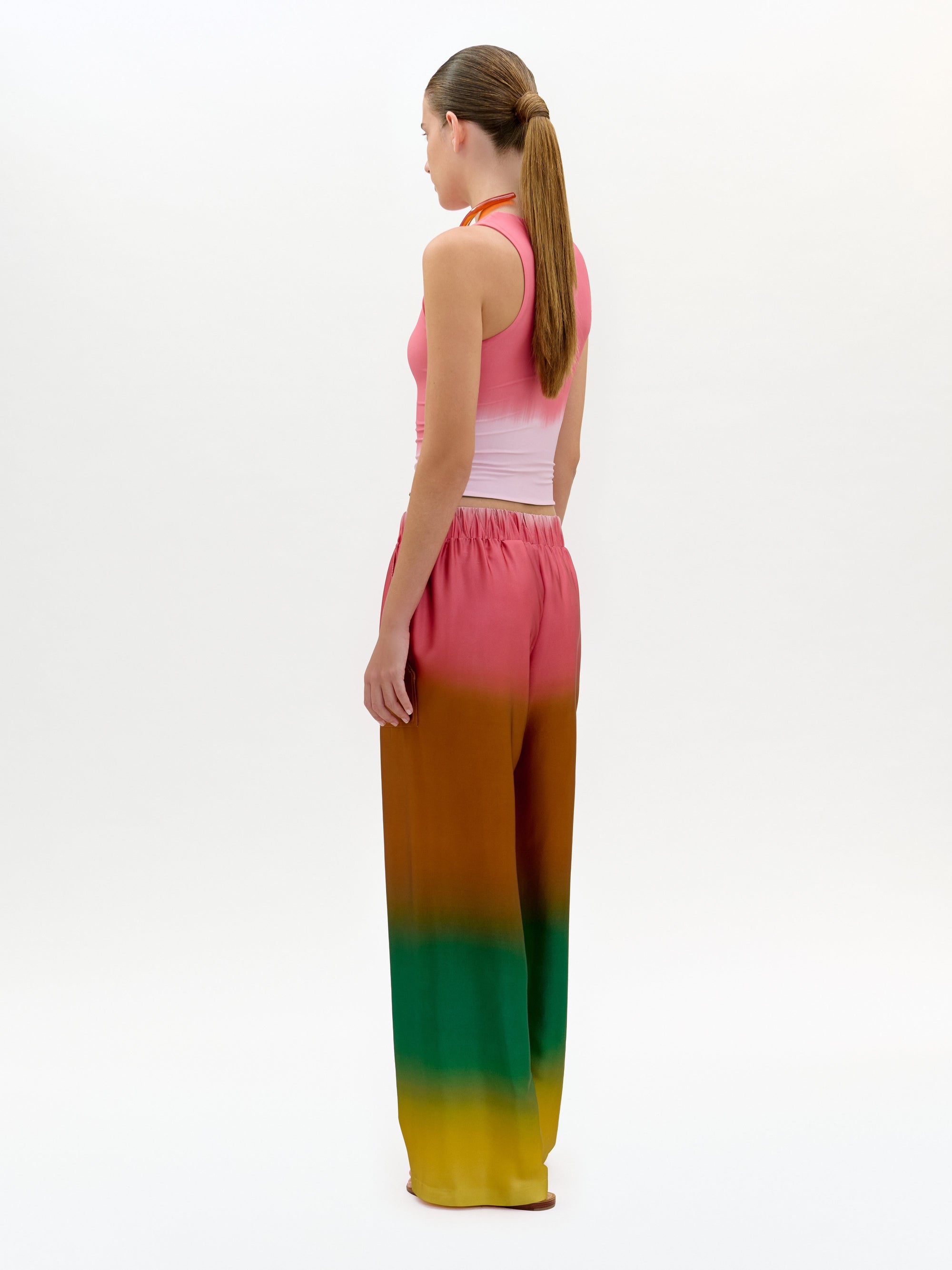 Una persona con el pelo largo recogido en una coleta baja, de pie y de lado, lleva un top rosa sin mangas y el pantalón Nerea -de talle alto y pernera ancha, con un efecto ombré que va del rosa al naranja y al verde- sobre un fondo blanco liso.