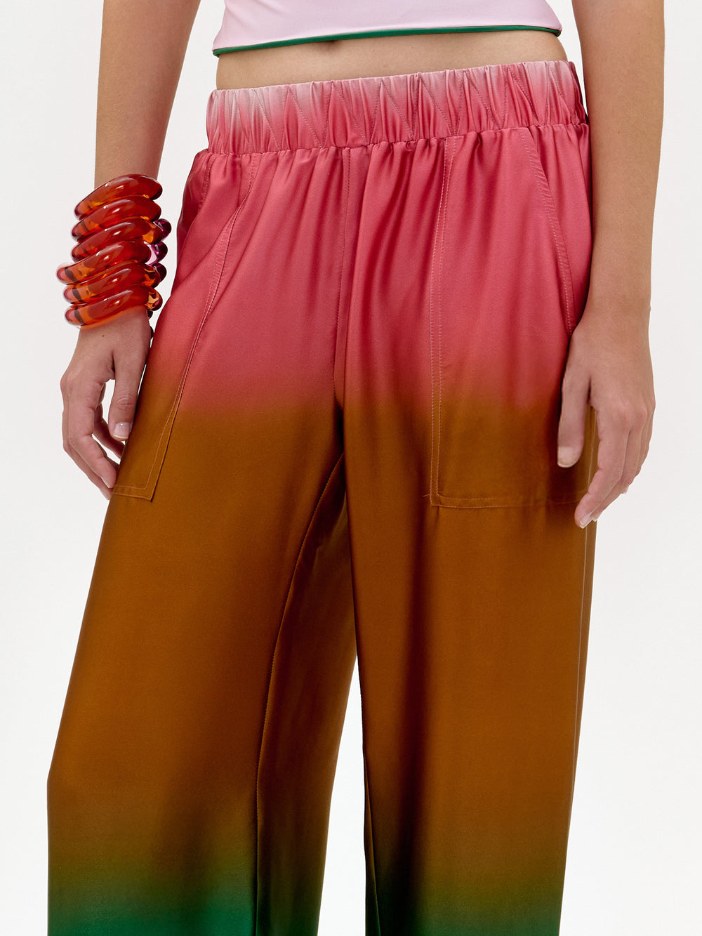 Una persona luce el pantalón Nerea, un pantalón de talle alto y pierna ancha fabricado en Colombia con un degradado ombré de rosa a naranja, combinado con un crop top blanco y un llamativo brazalete rojo sobre un fondo blanco liso.